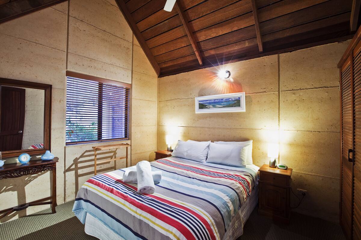 A bedroom with a wooden ceiling, a bed with a striped bedspread, a bedside table with a lamp, a window with blinds, and a framed picture on the wall.
