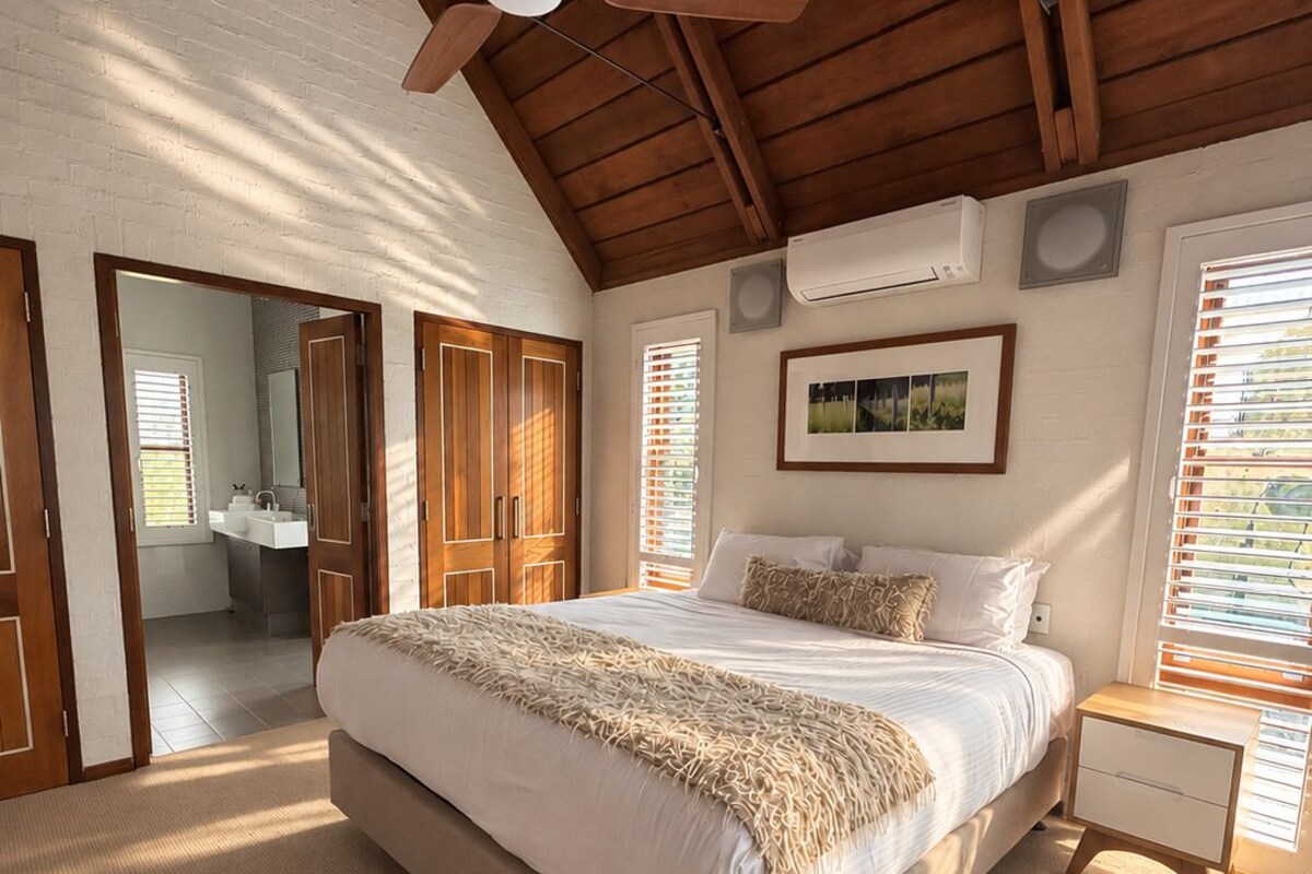 A bedroom with a large bed, wooden ceiling beams, a ceiling fan, and a window with blinds.