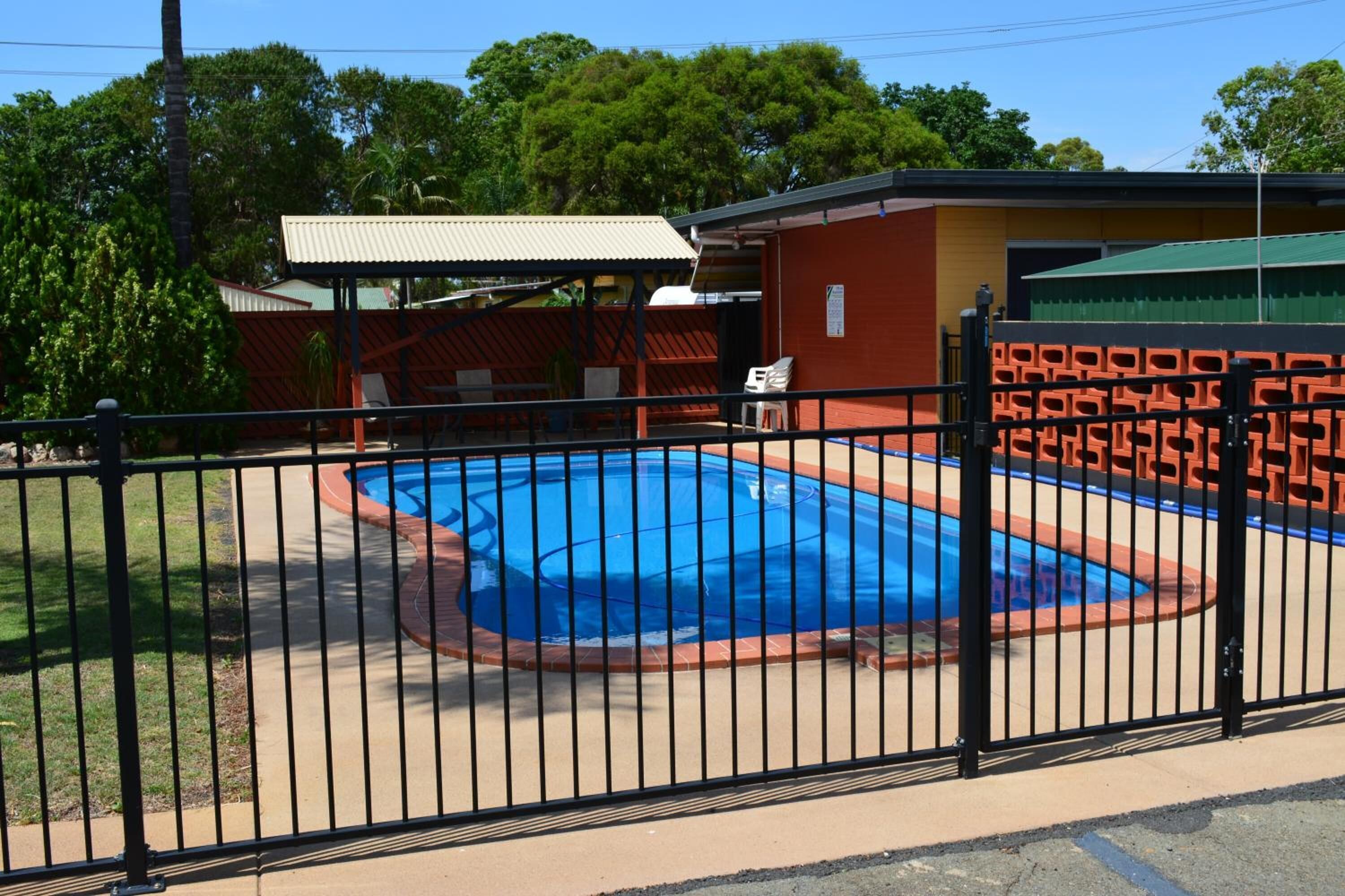 Ein von einer Umzäunung umschlossener Poolbereich mit überdachter Sitzgelegenheit und einem Gebäude im Hintergrund.