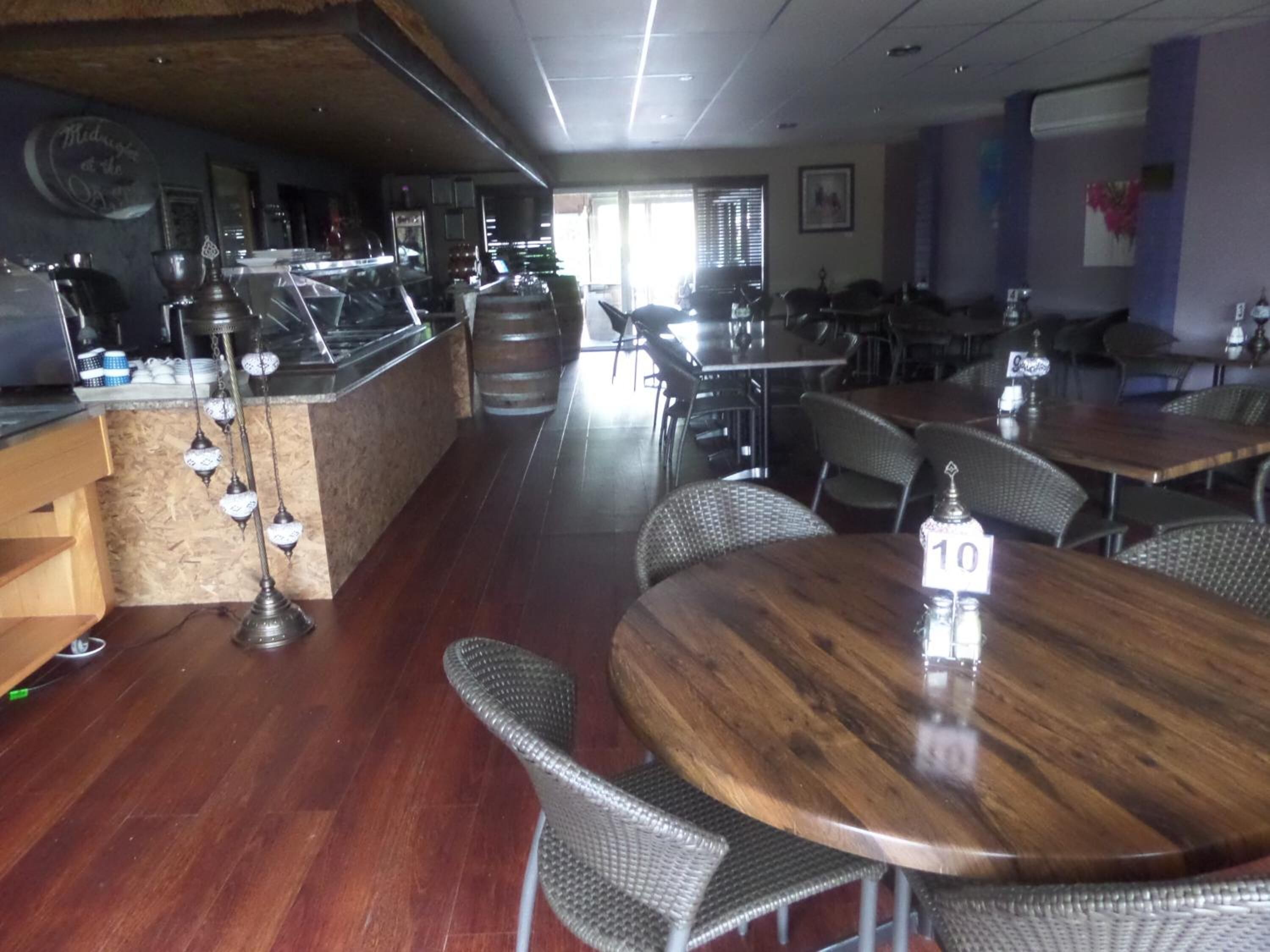 Un interior de restaurante con mesas y sillas de madera, una zona de bar con taburetes y vistas a la calle a través de amplias ventanas.