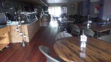 Un interior de restaurante con mesas y sillas de madera, una zona de bar con taburetes y vistas a la calle a través de amplias ventanas.