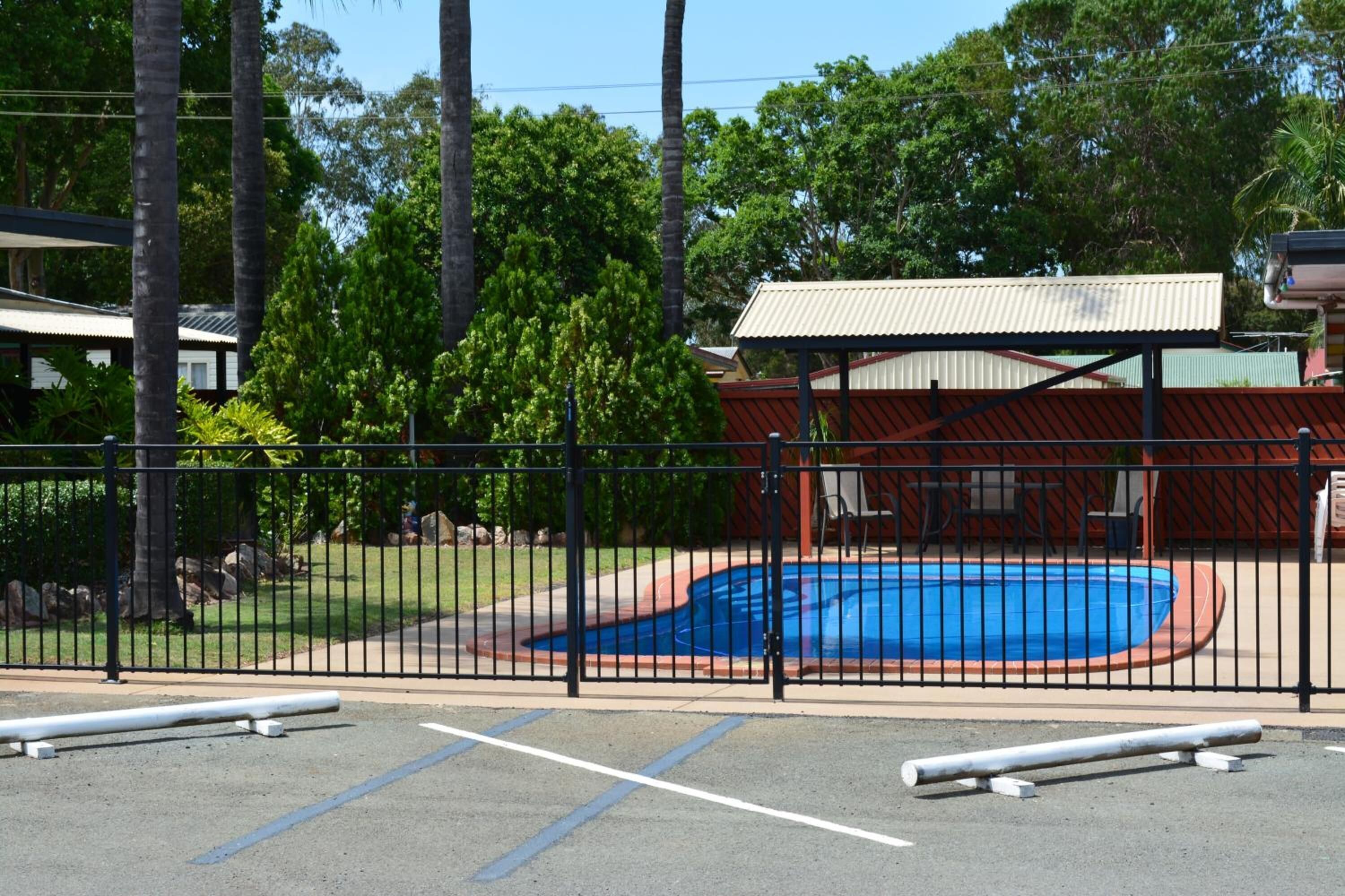 A fenced pool area with a blue pool, surrounded by trees and a building with a red roof.