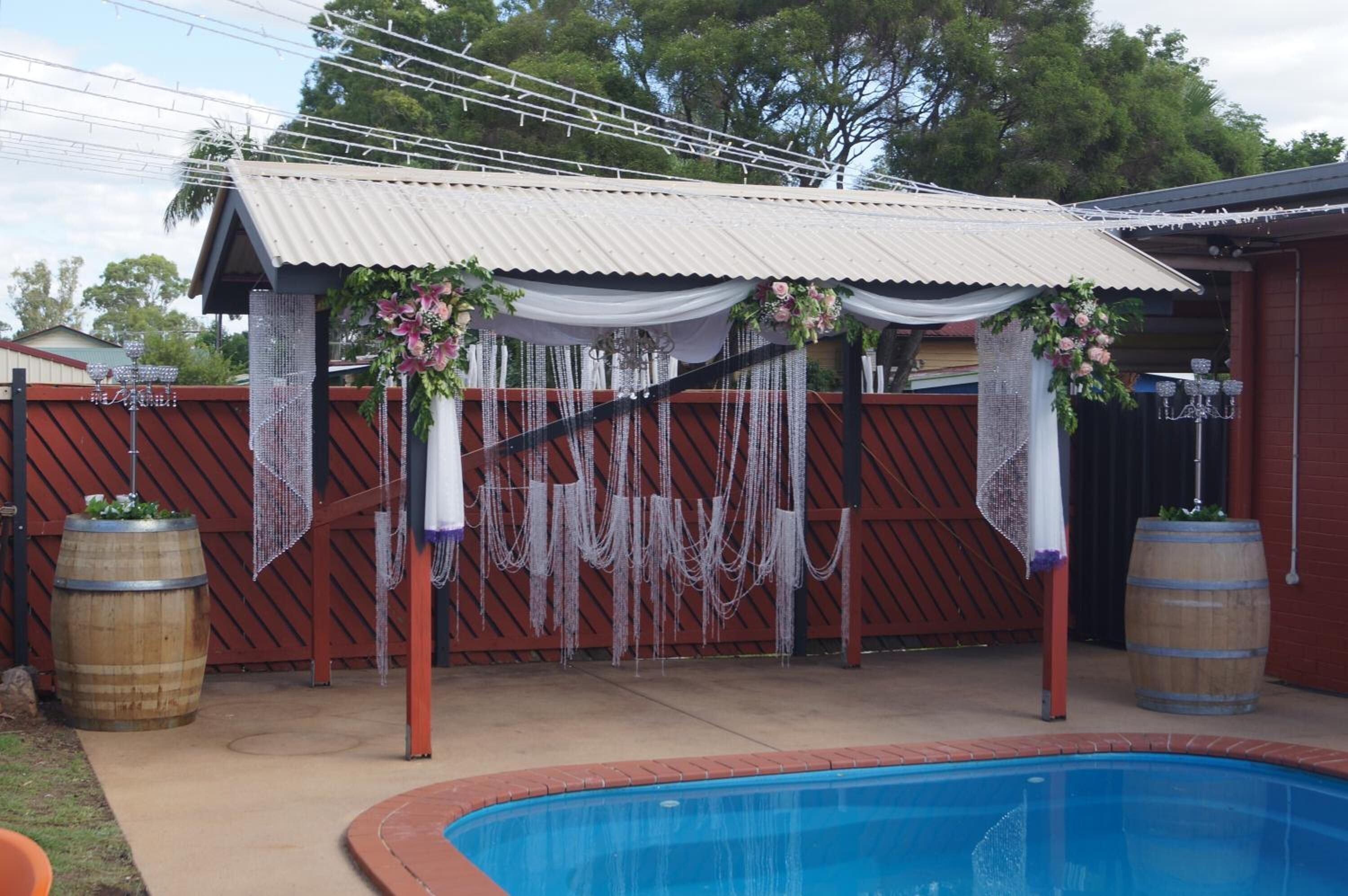 A backyard with a swimming pool, a covered patio area with draped fabric and hanging decorations, and wooden barrels.