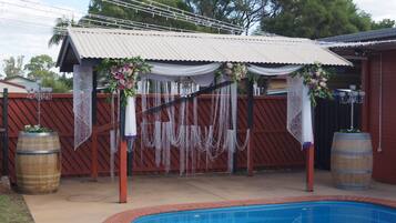 A backyard with a swimming pool, a covered patio area with draped fabric and hanging decorations, and wooden barrels.