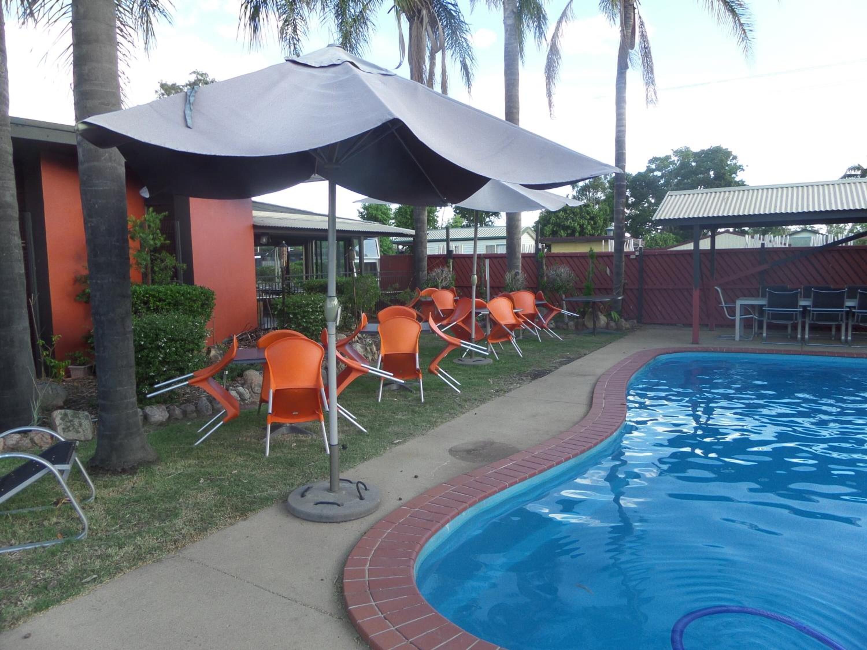 Una piscina con borde de ladrillo, rodeada de sillas y sombrillas naranjas, y palmeras al fondo.