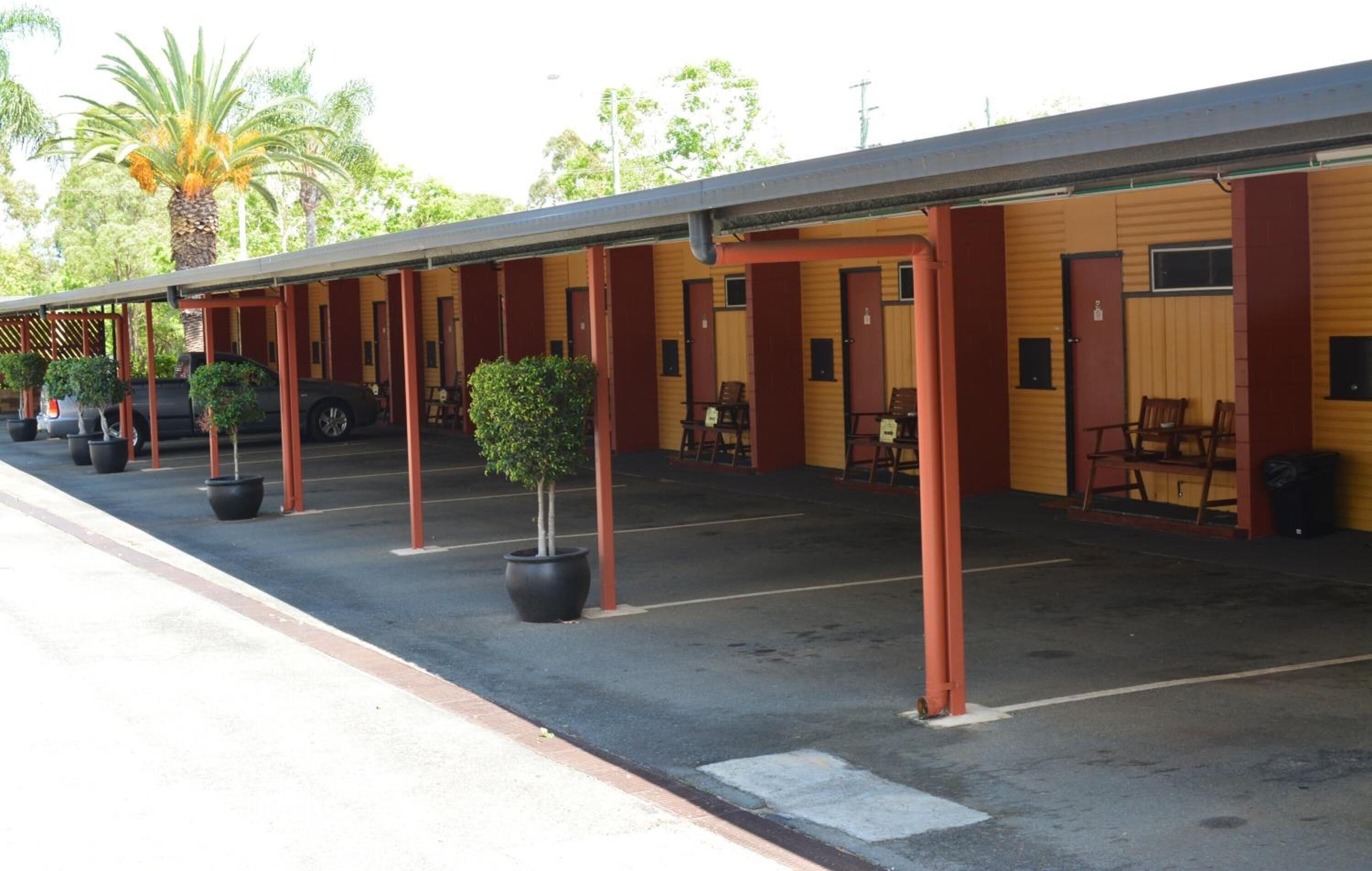 A motel with multiple rooms, each with a door and window, and a covered outdoor area with chairs and potted plants.