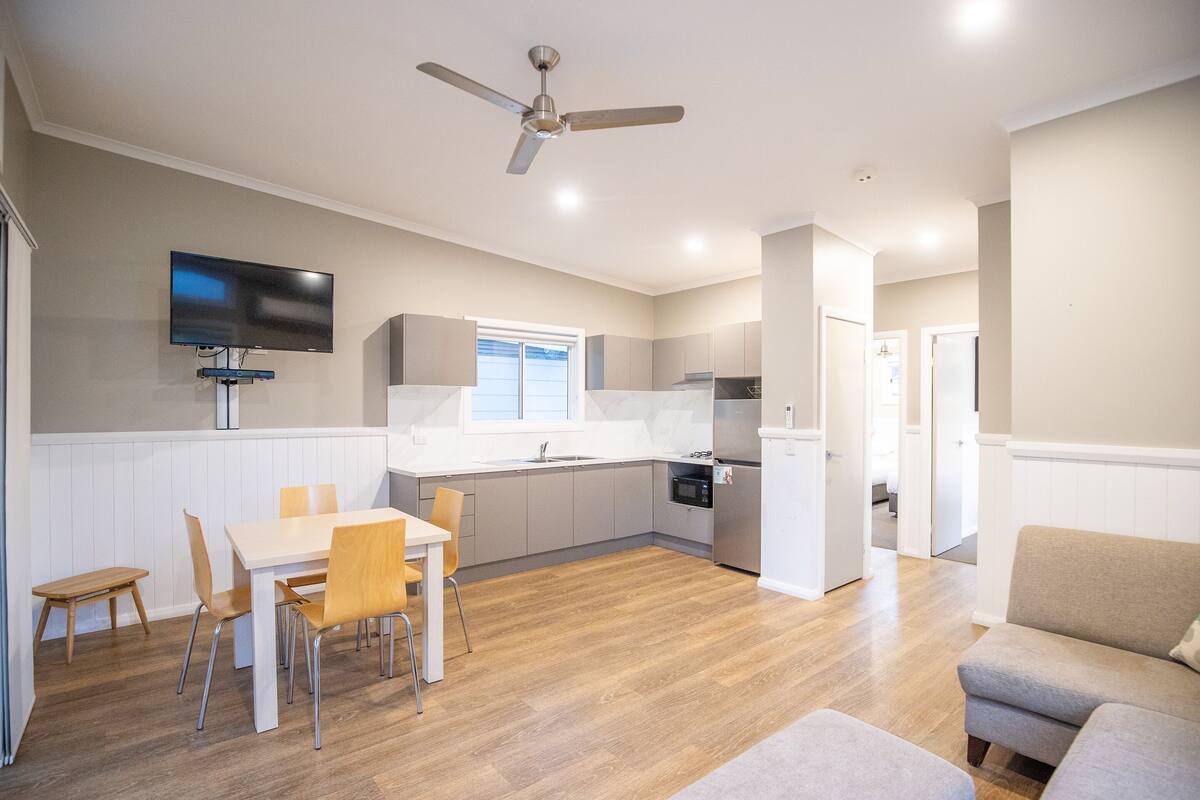 A modern kitchen with a flat-screen TV, wooden flooring, and a dining area with a table and chairs.