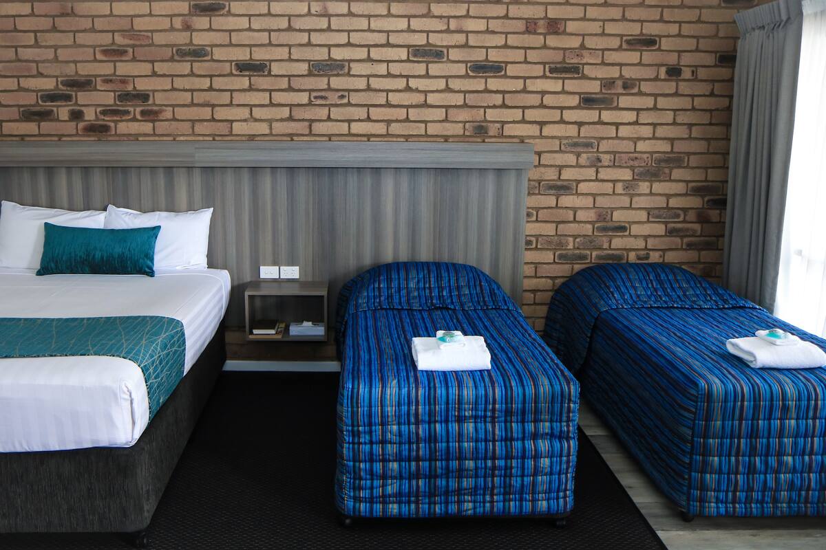 Una habitación de hotel con pared de ladrillo, una cama con ropa blanca y una manta verde azulada, dos camas individuales con ropa azul, y una pequeña mesa entre ellas.
