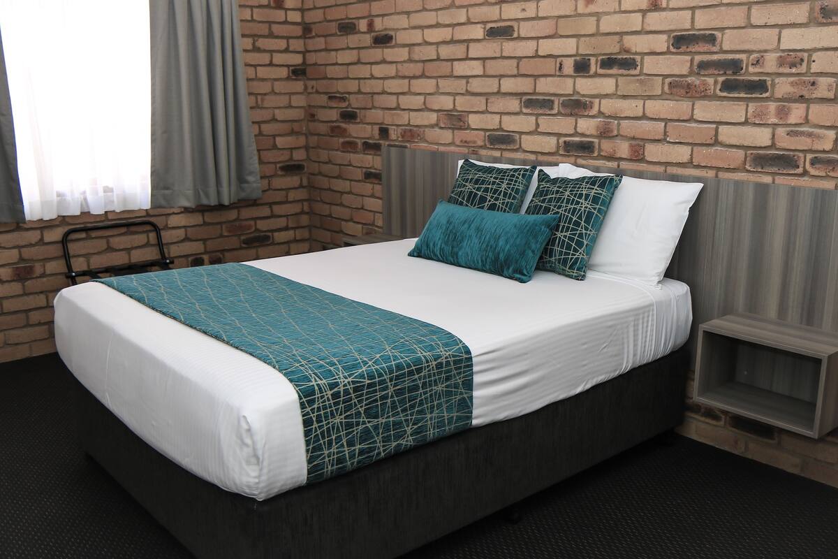 Una cama con una colcha blanca y azul verdosa, dos almohadas y un cabecero frente a una pared de ladrillo.