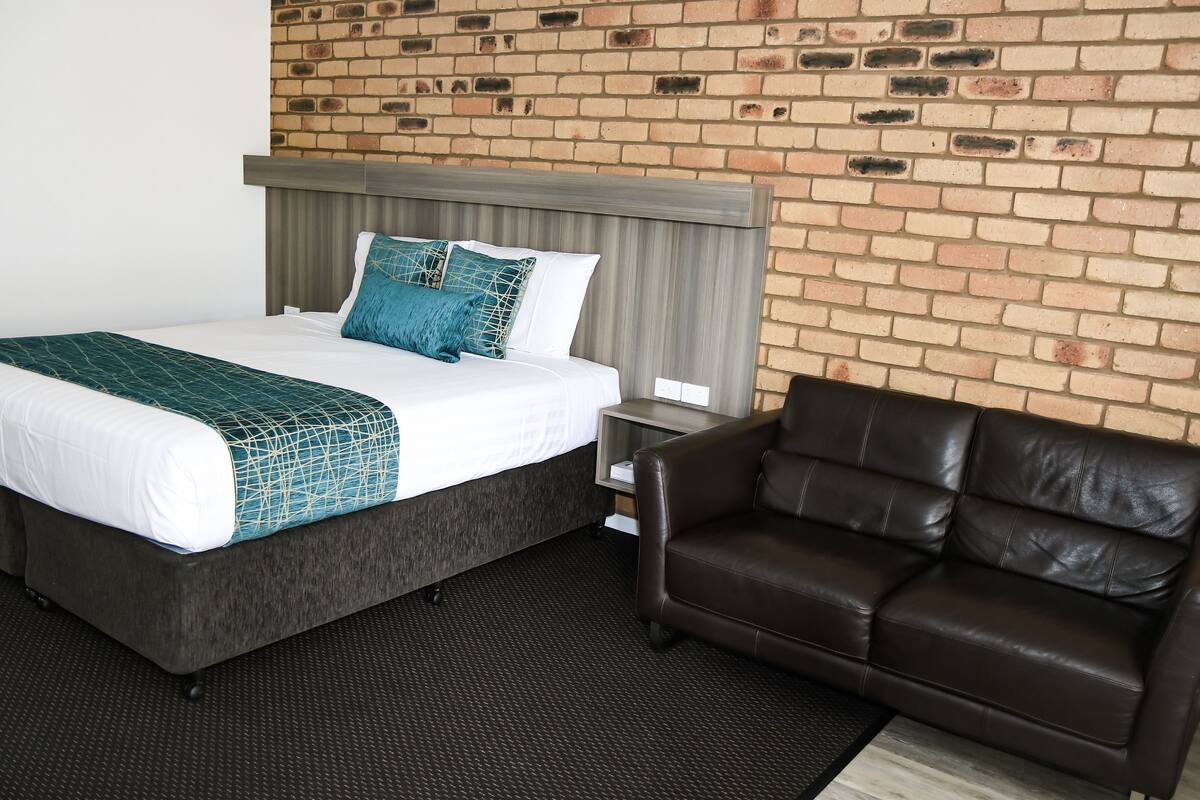 Una habitación de hotel con una cama, un sofá de cuero marrón, una pared de ladrillo y un cabecero de madera.