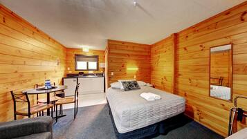 A wooden cabin room with a bed, a small table, a chair, a desk, and a mirror.