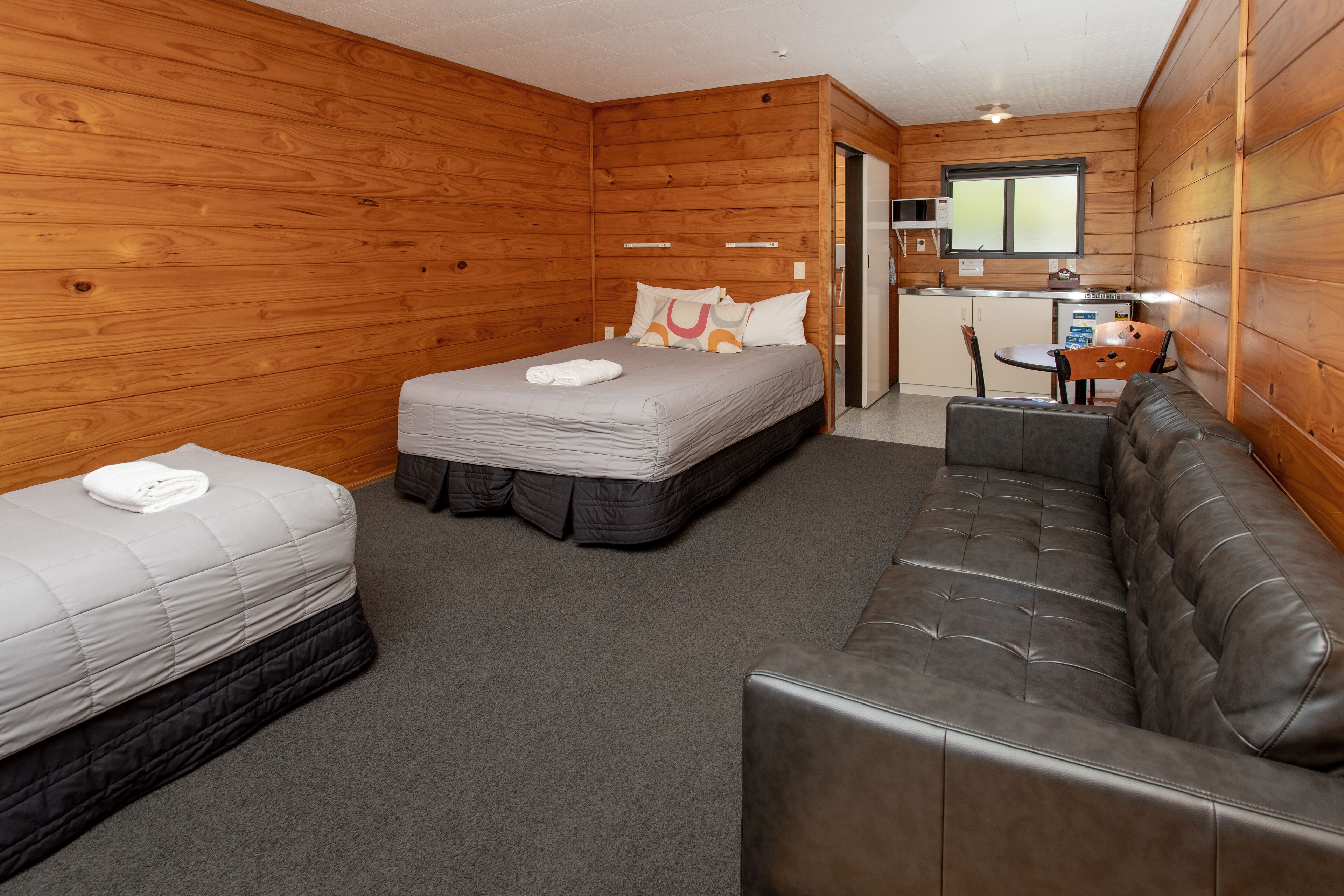 A room with two beds, a sofa, a small kitchen area, and wooden walls.