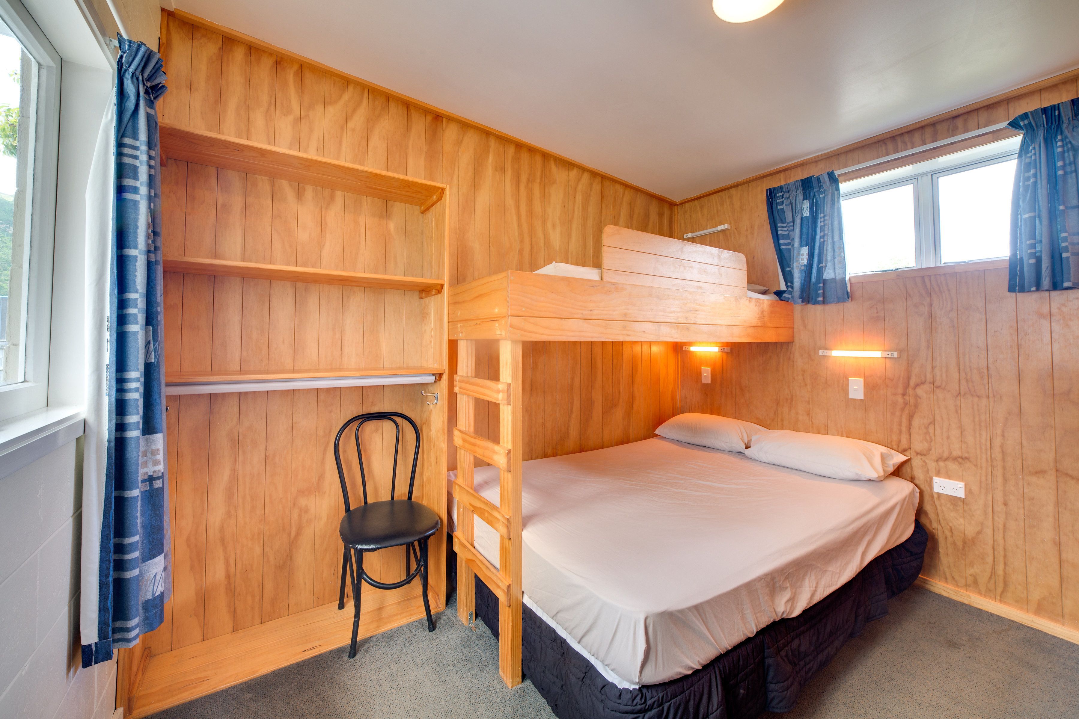 A wooden bunk bed room with a desk and chair, a window with curtains, and a wall-mounted light fixture.
