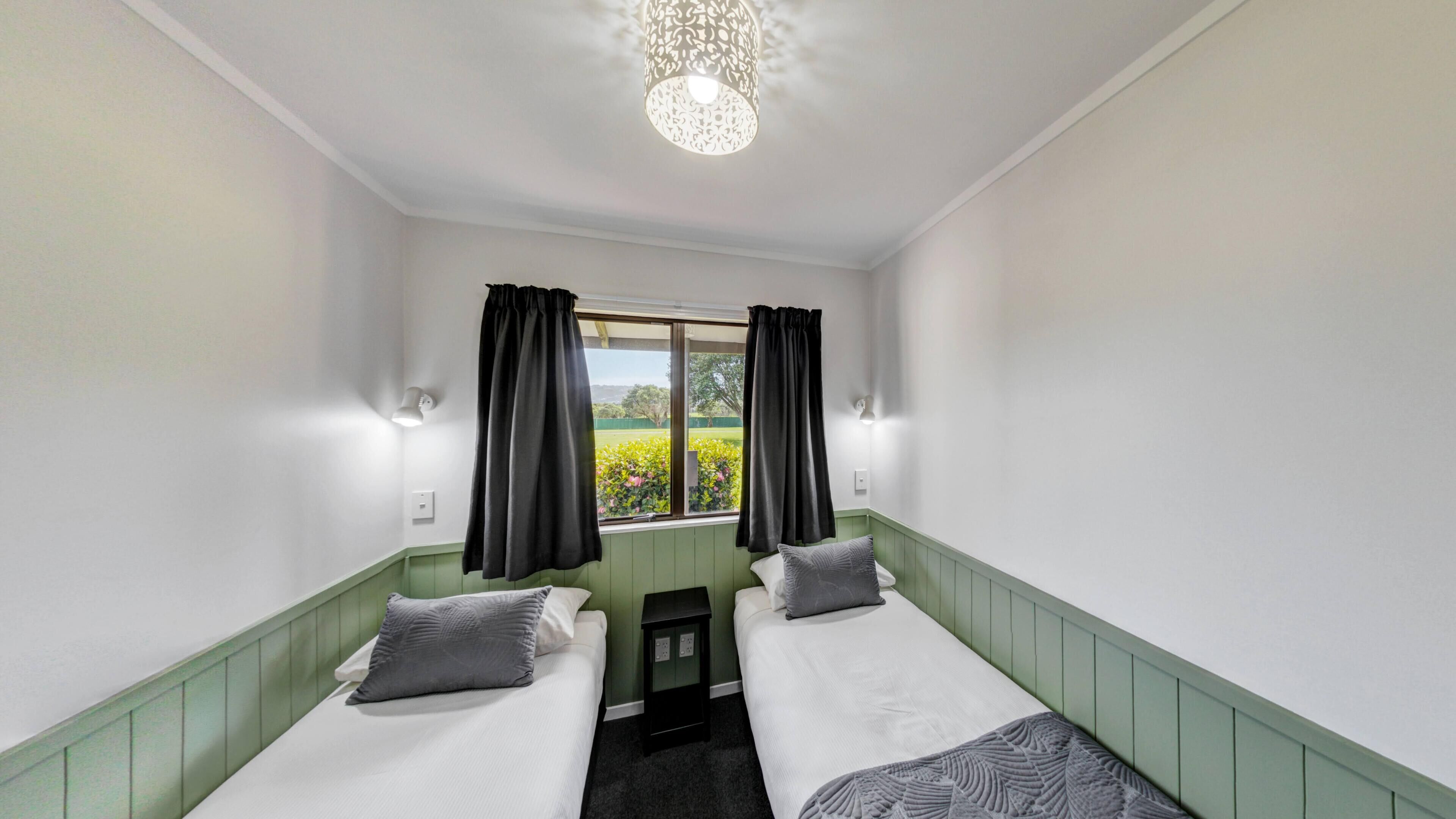 A room with two beds, a window with curtains, a chandelier, and a view of greenery outside.