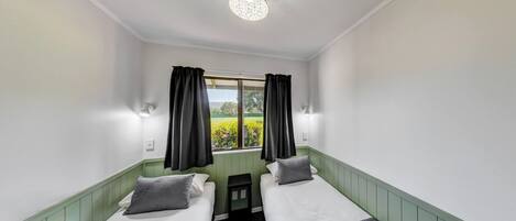 A room with two beds, a window with curtains, a chandelier, and a view of greenery outside.