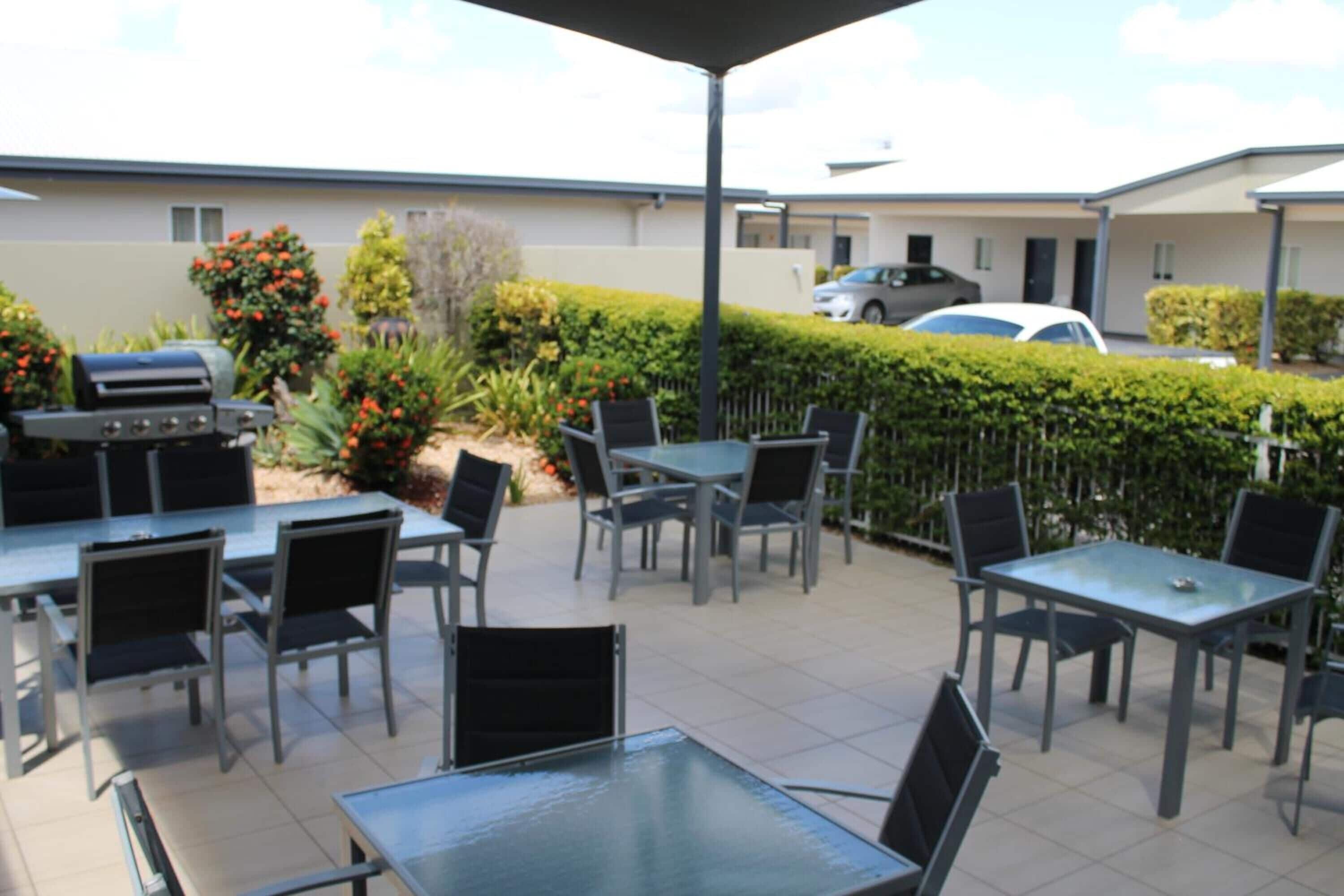 Eine Terrasse im Freien mit Tischen und Stühlen, einem Grill und einem Hecken im Hintergrund.