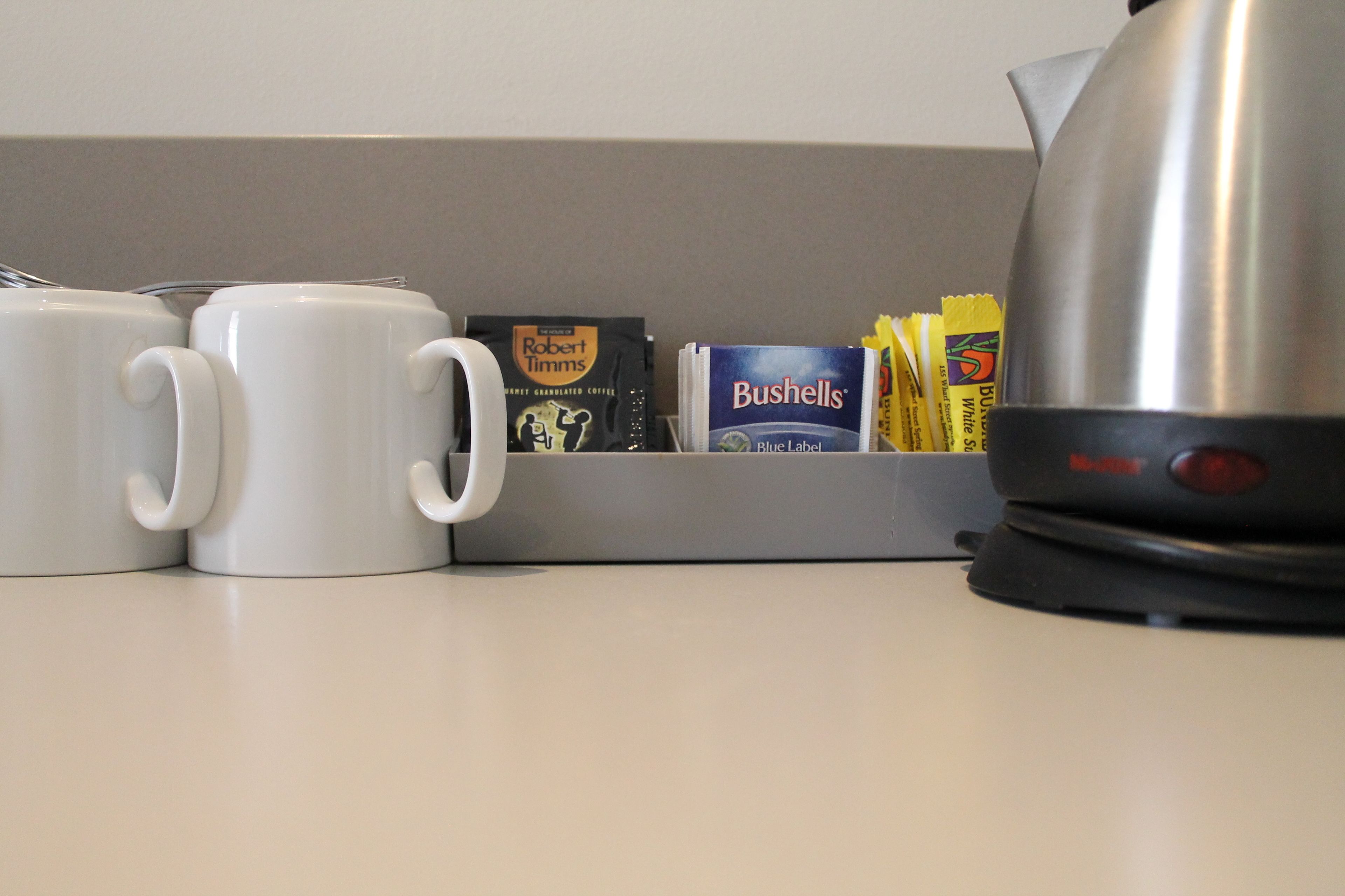 Una encimera de cocina con dos tazas blancas, una cafetera y varios productos de café, incluyendo un paquete de café Robert Timms y una caja de café Bushells.