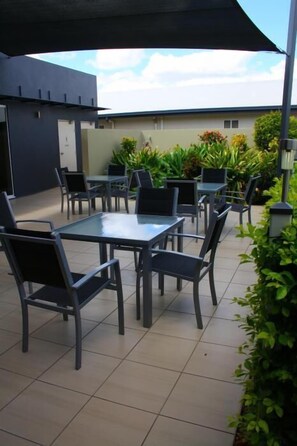 Un patio al aire libre con varias mesas y sillas, rodeado de plantas y un edificio al fondo.