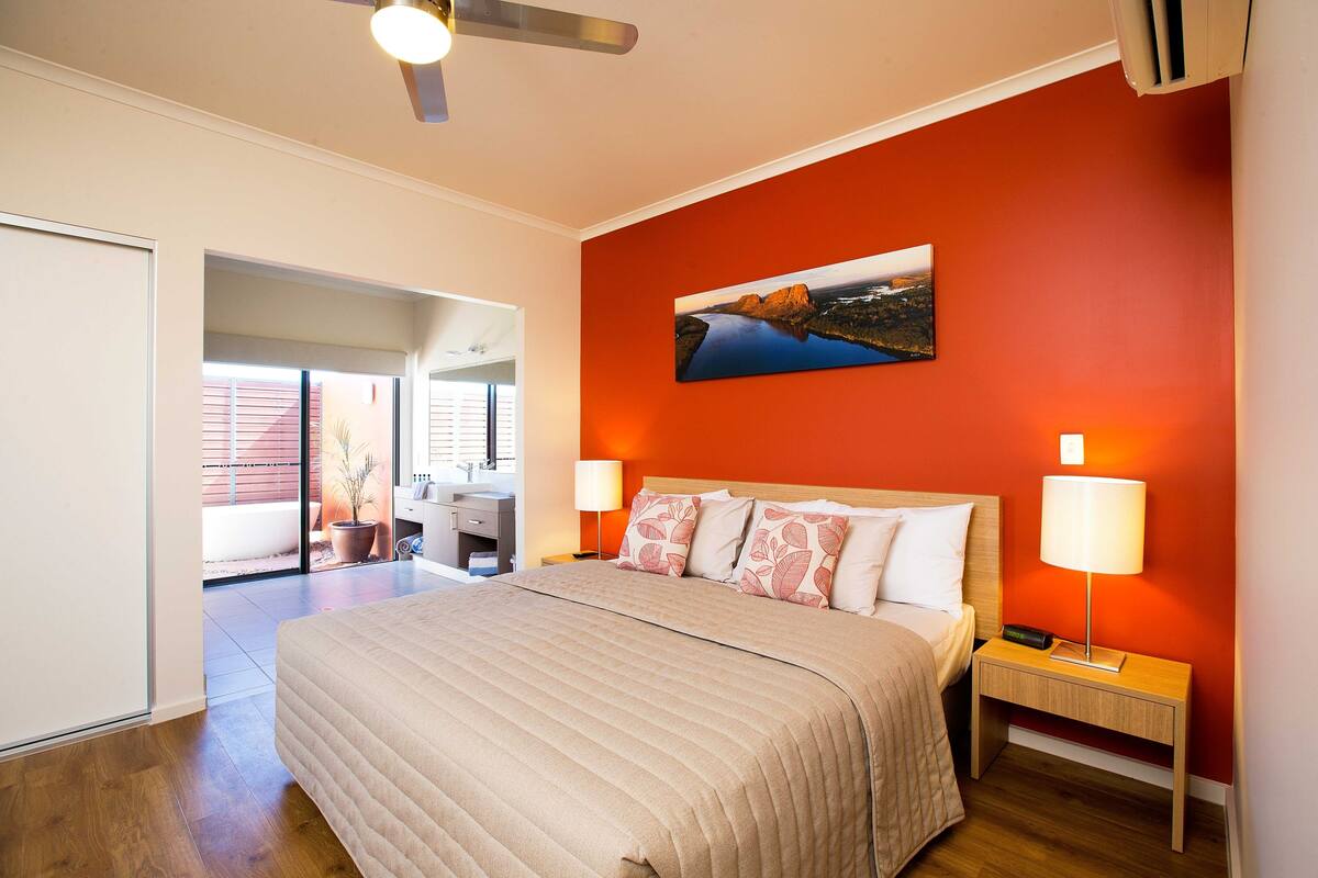Une chambre à coucher avec un grand lit, une table de chevet avec une lampe, un ventilateur de plafond et un tableau au mur.