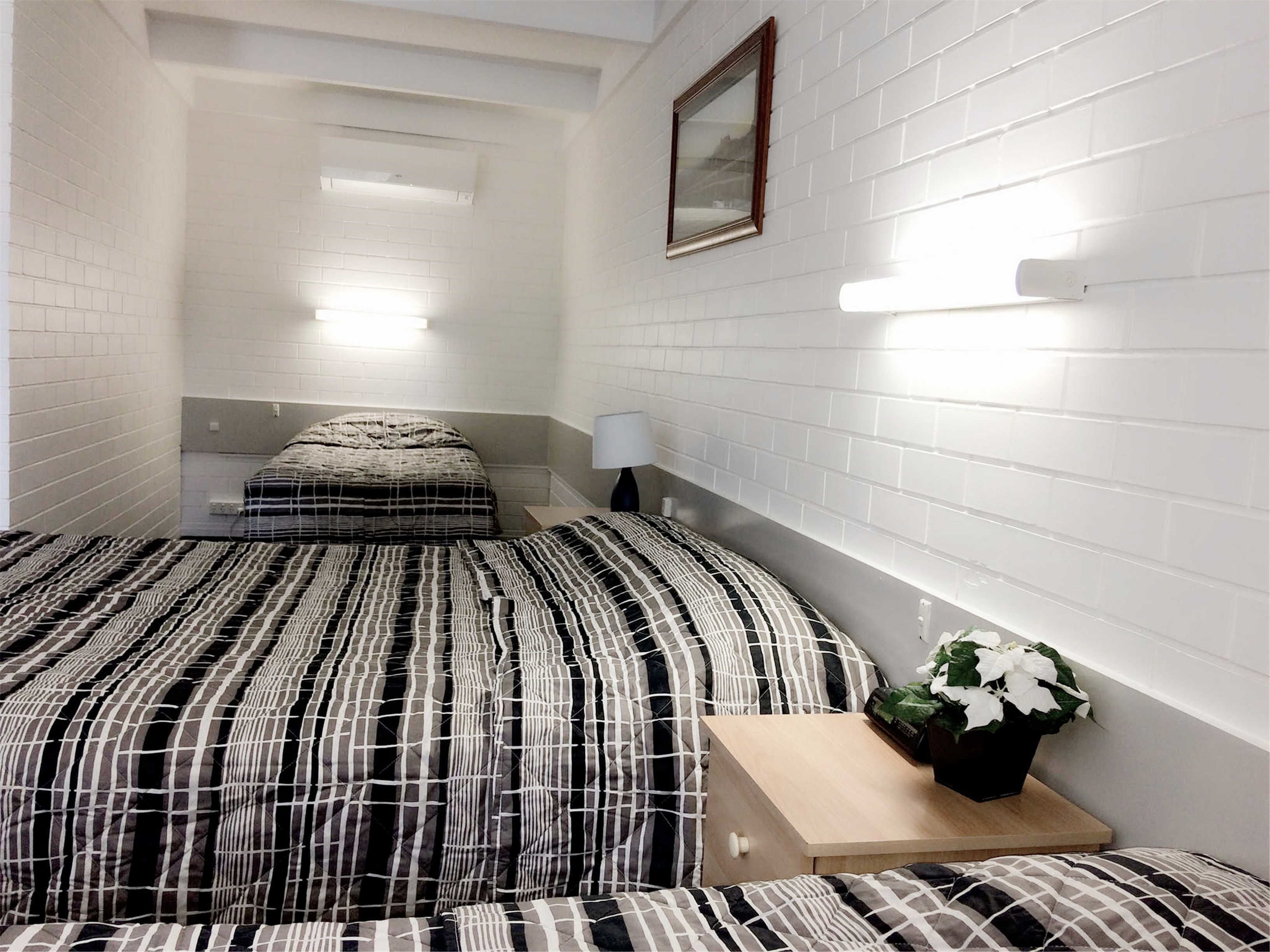 Ein kleiner, schmaler Raum mit weißen Ziegelwänden, einem Bett mit gestreifter Bettdecke, einem Nachttisch mit Lampe und einem kleinen Topfpflanzen.