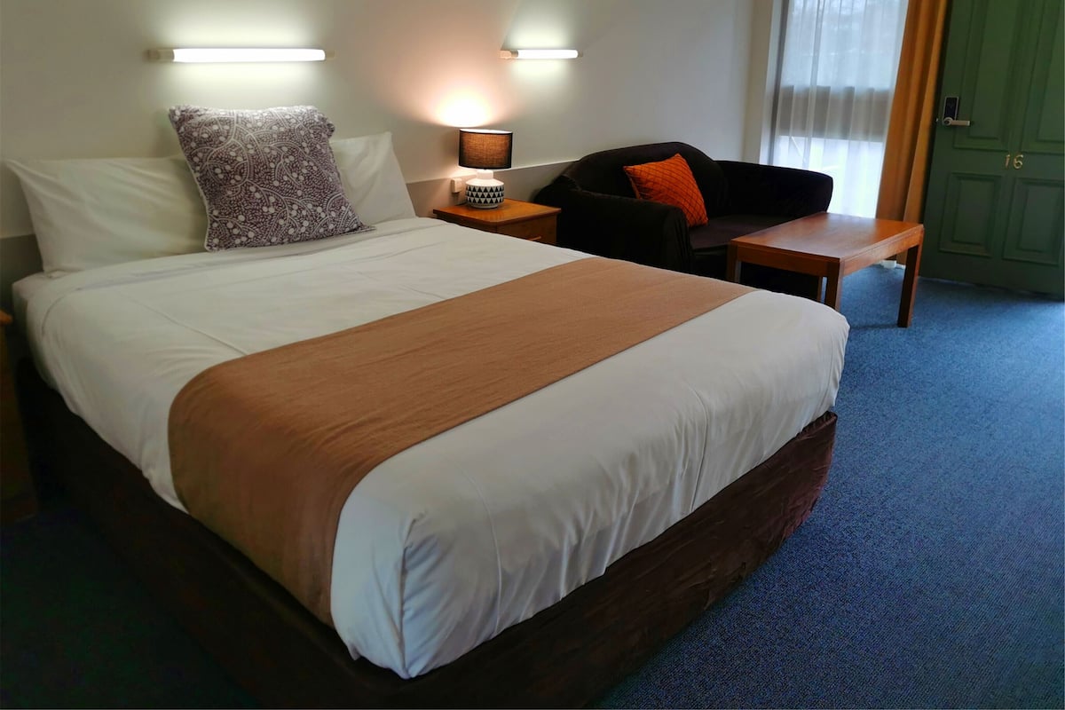 A hotel room with a large bed, a nightstand with a lamp, a chair, and a small table.