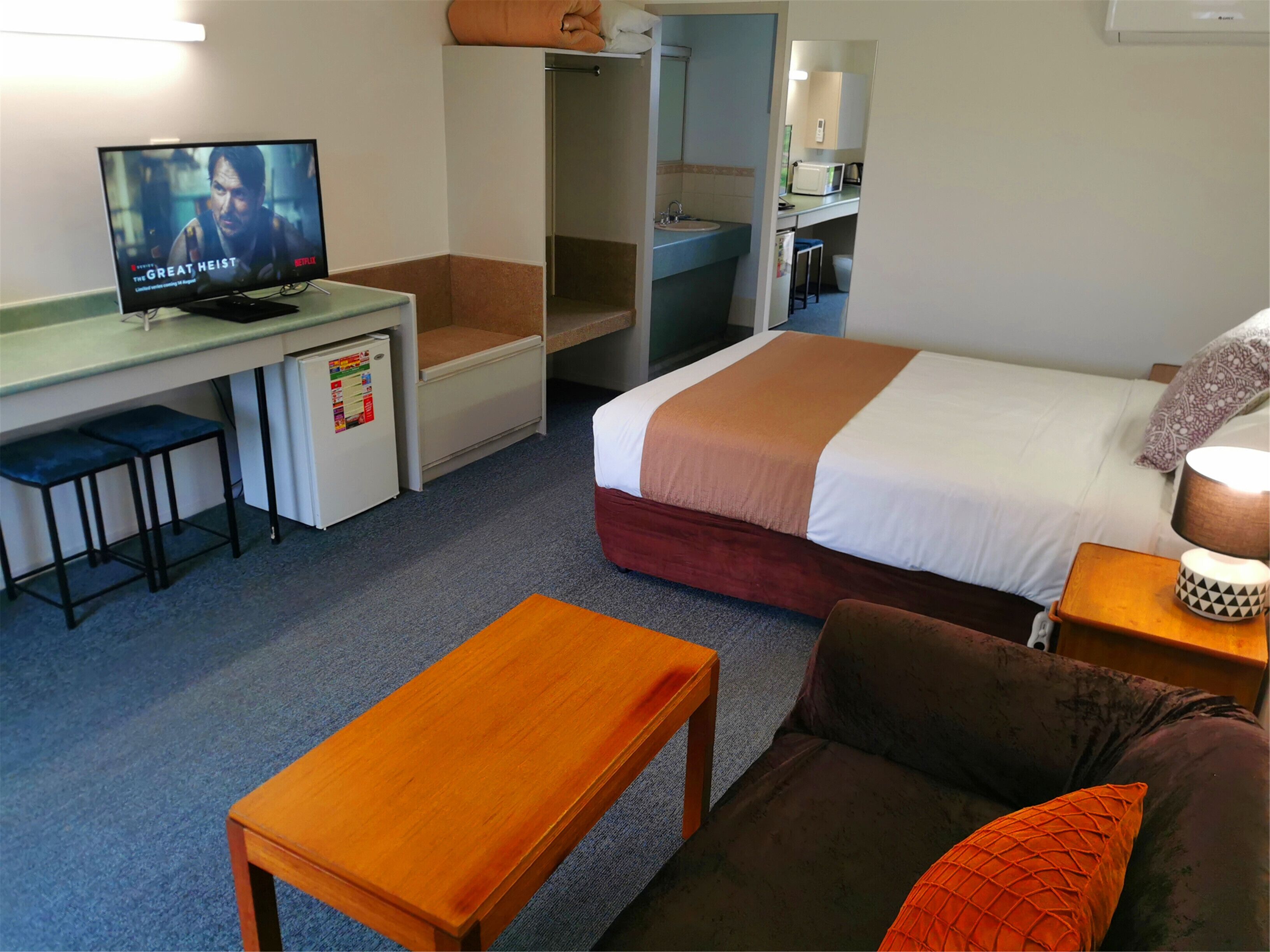 Habitación de hotel con una cama, un escritorio con televisor, un refrigerador pequeño, un banco de madera y un sofá.