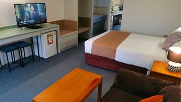 Habitación de hotel con una cama, un escritorio con televisor, un refrigerador pequeño, un banco de madera y un sofá.