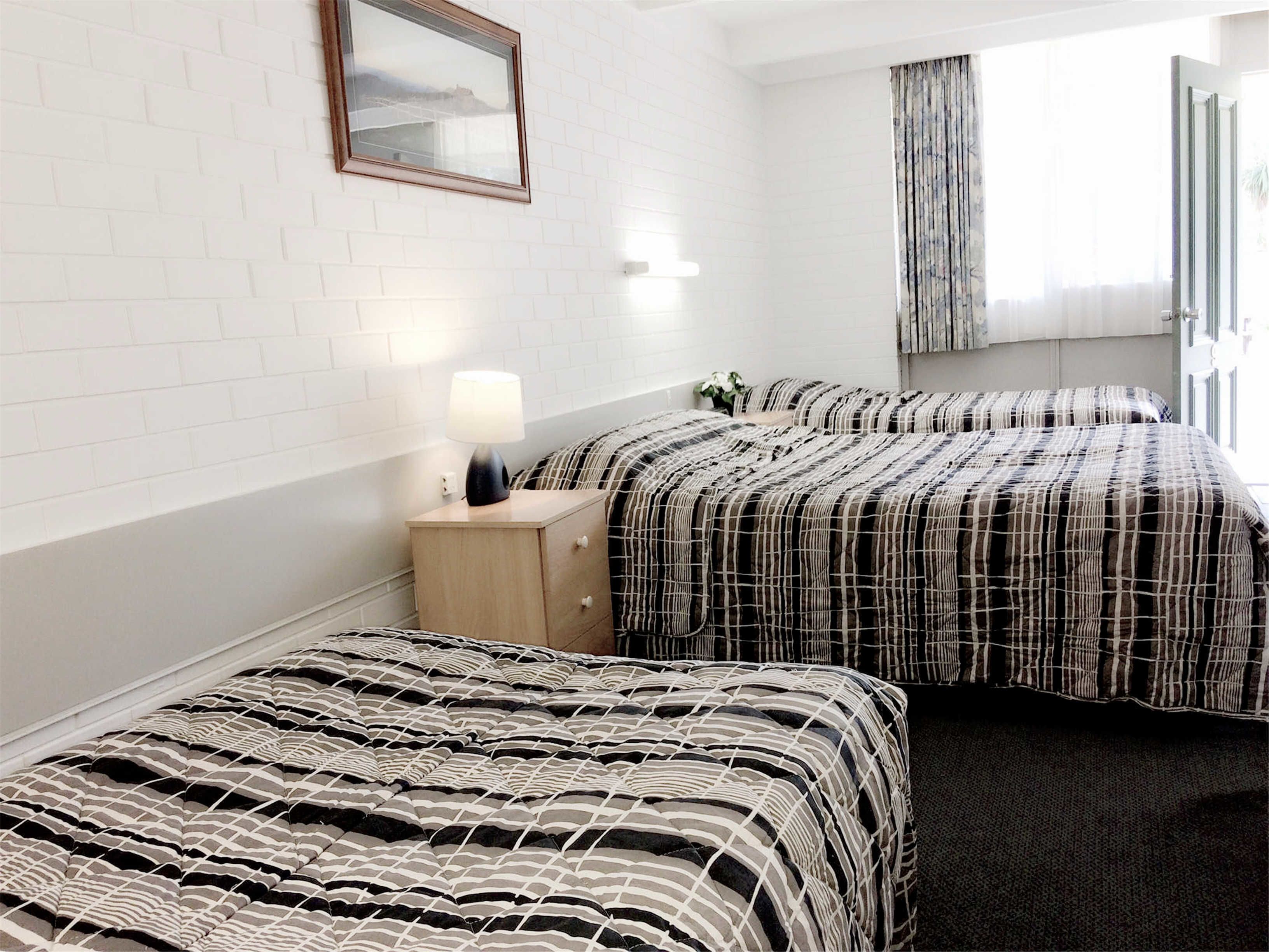 A hotel room with two beds, a nightstand with a lamp, a window with curtains, and a mirror on the wall.