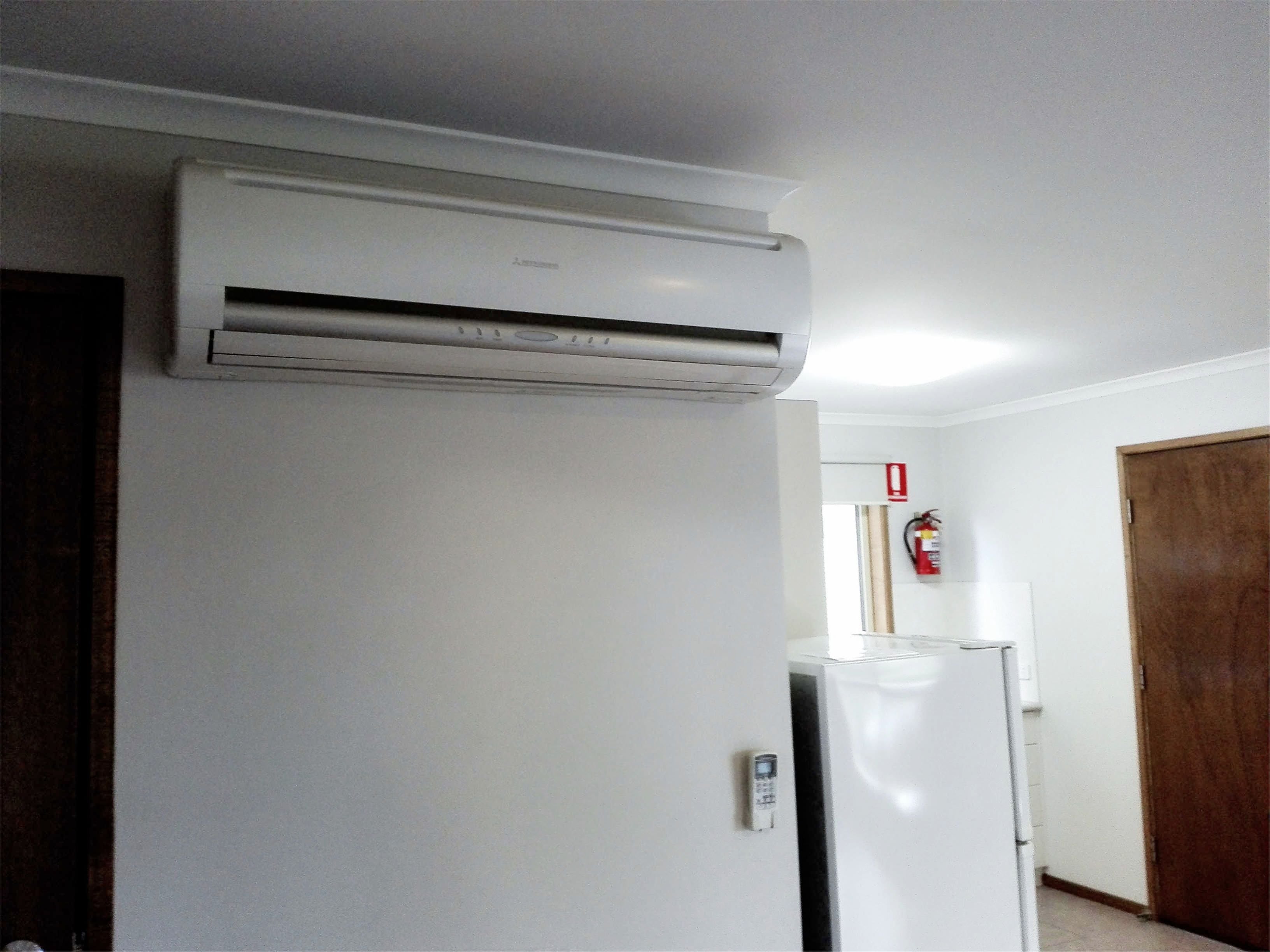 Un aparell d'aire condicionat blanc fixat a la paret, un frigorífic blanc i una porta de fusta en una habitació amb parets blanques.