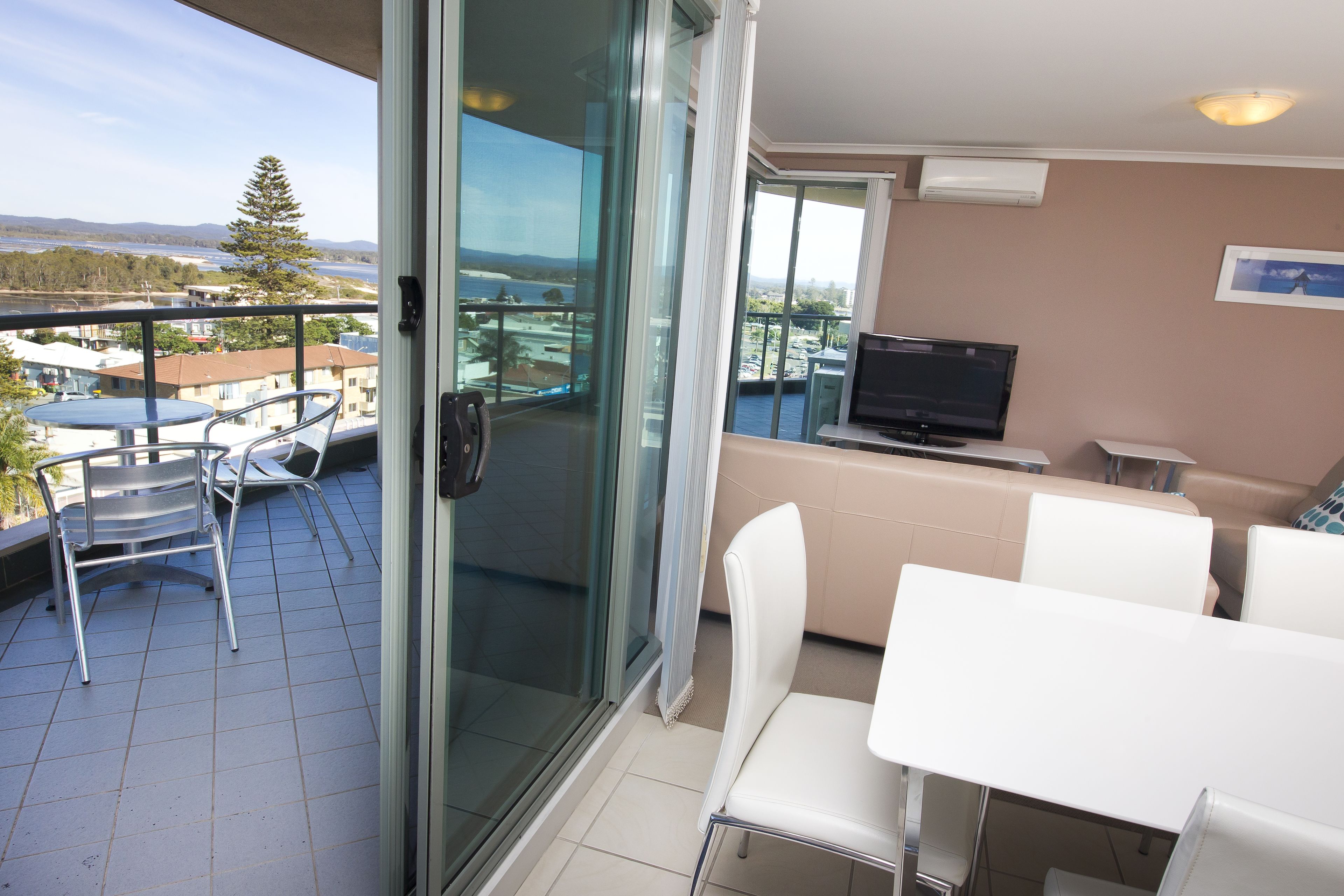 A balcony with a glass door, a dining table with chairs, a TV, and a view of a body of water and buildings.