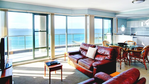 A spacious living room with a leather sofa, a small table, and a dining area with chairs. Large windows offer a view of the ocean. - Sails Luxury Apartments, Forster (Forster)