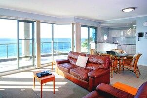 Uma sala de estar espaçosa com um sofá de couro, um espaço de jantar com cadeiras de vime e grandes portas de vidro deslizantes que dão para uma varanda com vista para o oceano.