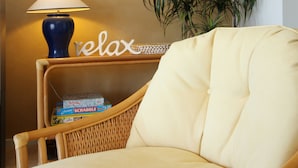 A wicker chair with a cushion, a lamp, a Scrabble box, and a plant. - Sails Luxury Apartments, Forster (Forster)