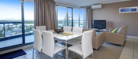 Una sala de estar moderna con una mesa de comedor, sillas blancas, un sofá, un televisor y ventanas grandes con cortinas.