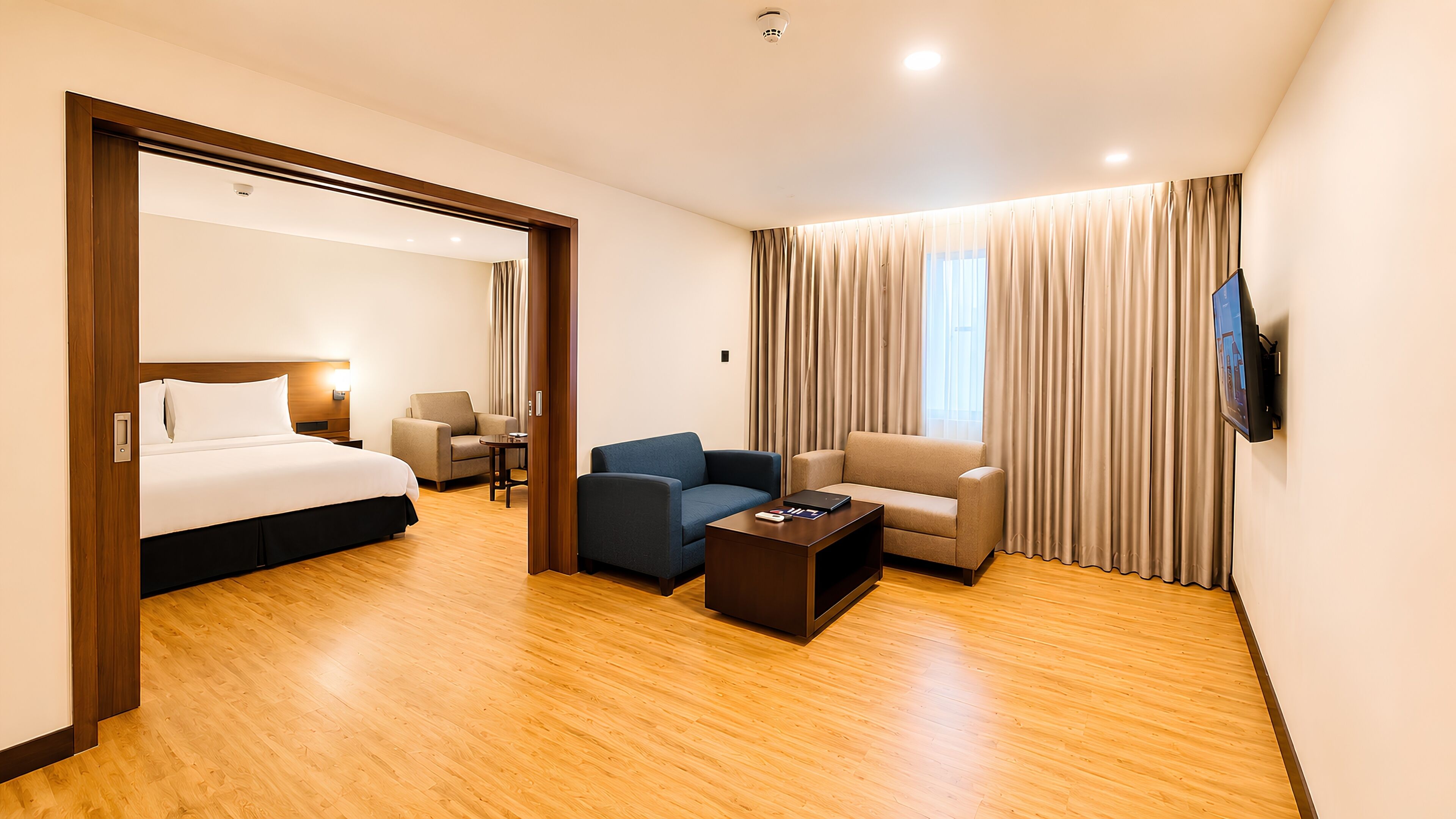A hotel room with a bed, two armchairs, a coffee table, a TV, and a large window with curtains.