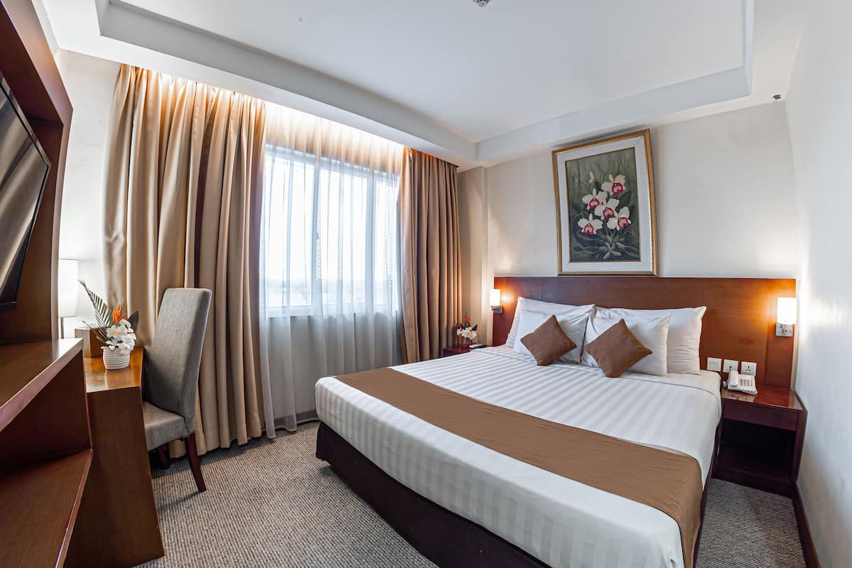 A hotel room with a large bed, a desk with a chair, a desk lamp, and a painting on the wall.