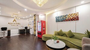 A modern hotel lobby with a reception desk, a Coca-Cola refrigerator, a green sofa, and a chandelier.