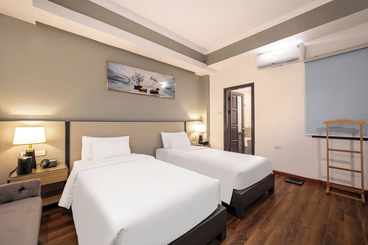 Een hotelkamer met twee bedden, een bureau met stoel, een bureaulamp, een schilderij aan de muur, een airconditioner en een houten ladder.