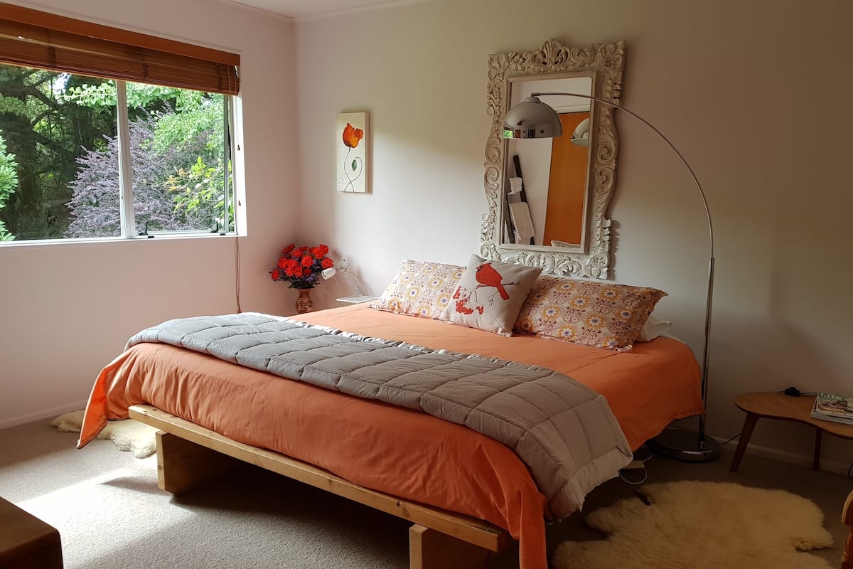 A bedroom with a bed, a nightstand, a lamp, a mirror, and a window with a view of trees.
