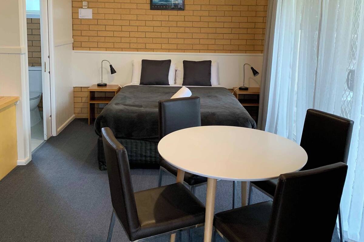 A compact hotel room with a bed, a small table, and two chairs. There is a brick wall with a picture, a window with curtains, and a ceiling light.