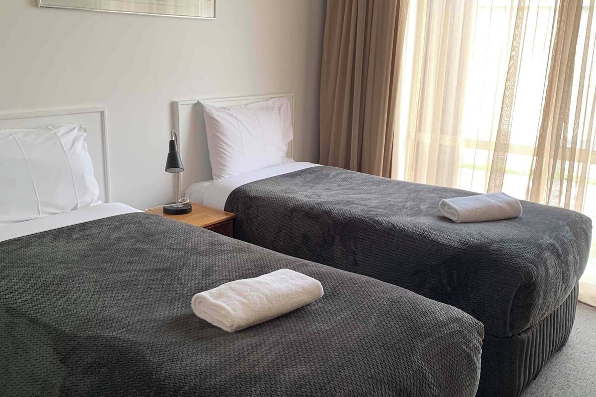 A hotel room with two beds, a nightstand, a lamp, and a window with curtains.