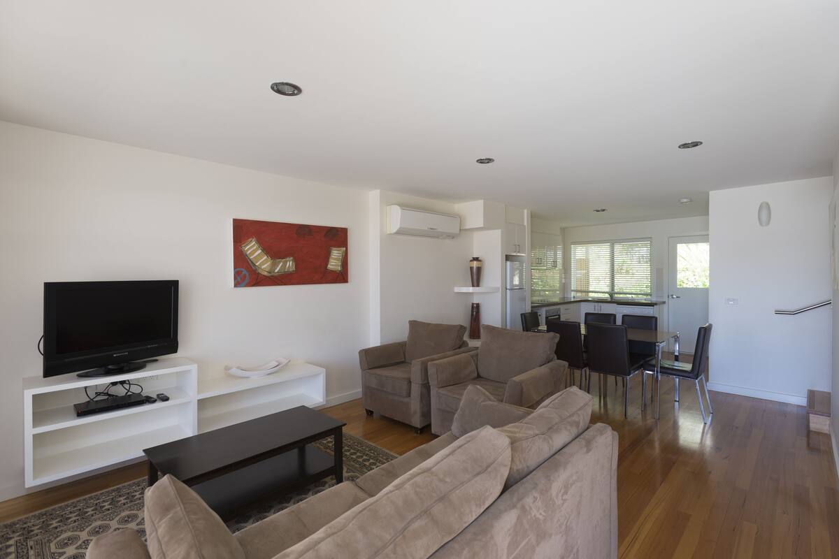 A modern living room with a flat-screen TV, a white entertainment unit, beige sofas, and a dining area with a table and chairs.