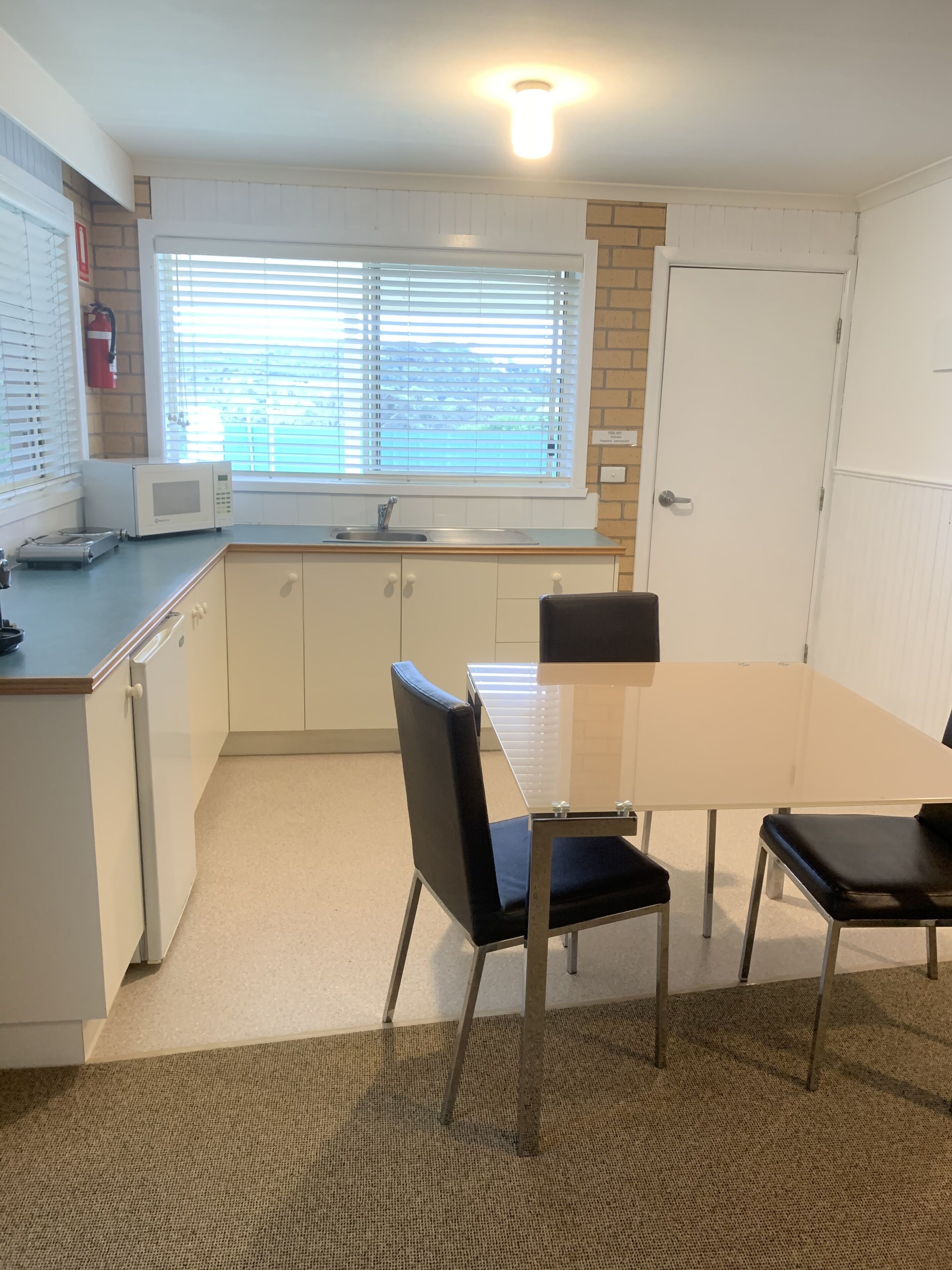 A compact kitchen with a microwave, sink, and window with blinds. There is a table with chairs and a rug on the floor.