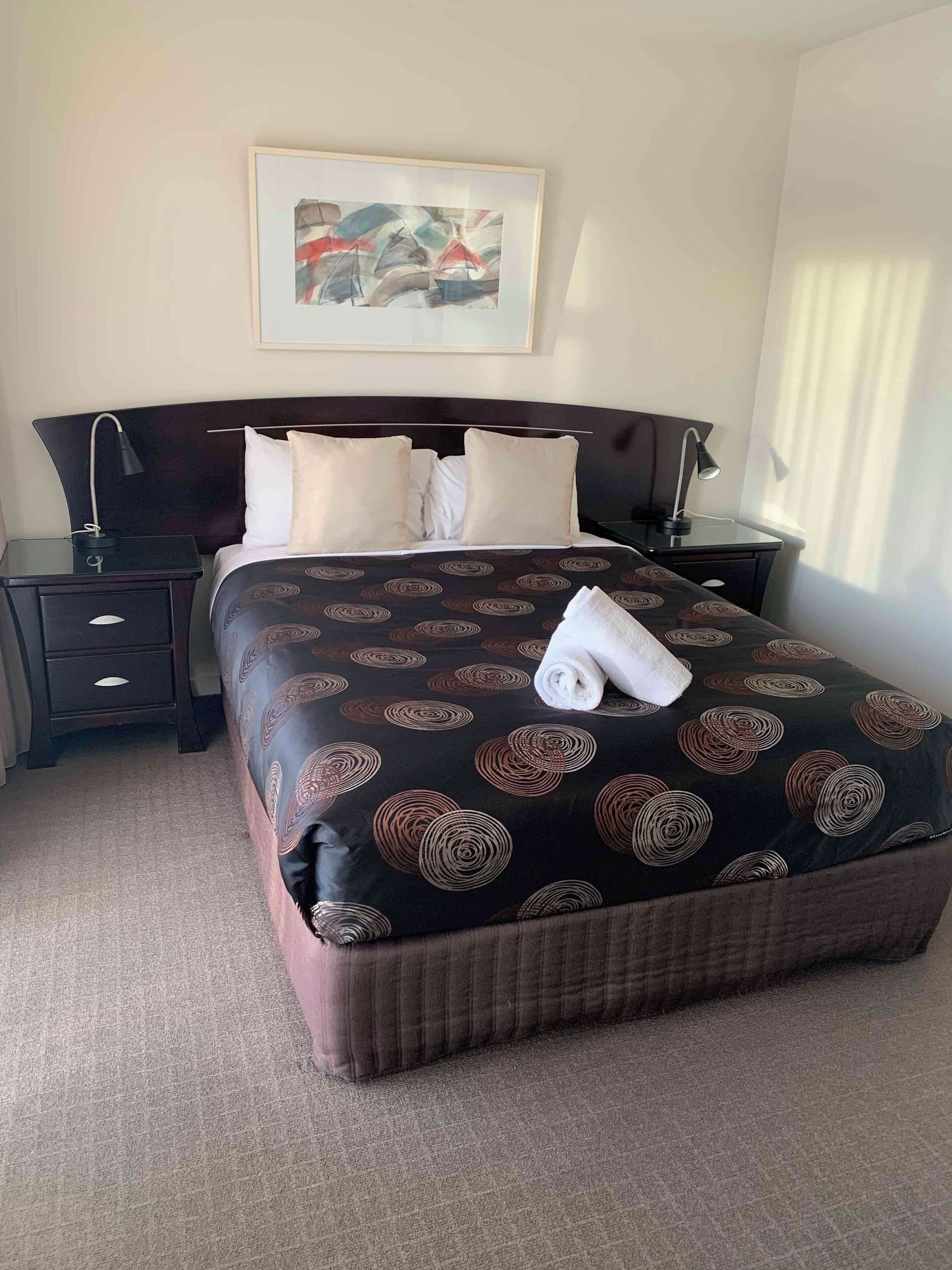 A neatly made bed with a patterned bedspread, two bedside tables with lamps, a wall-mounted artwork, and a window with curtains.