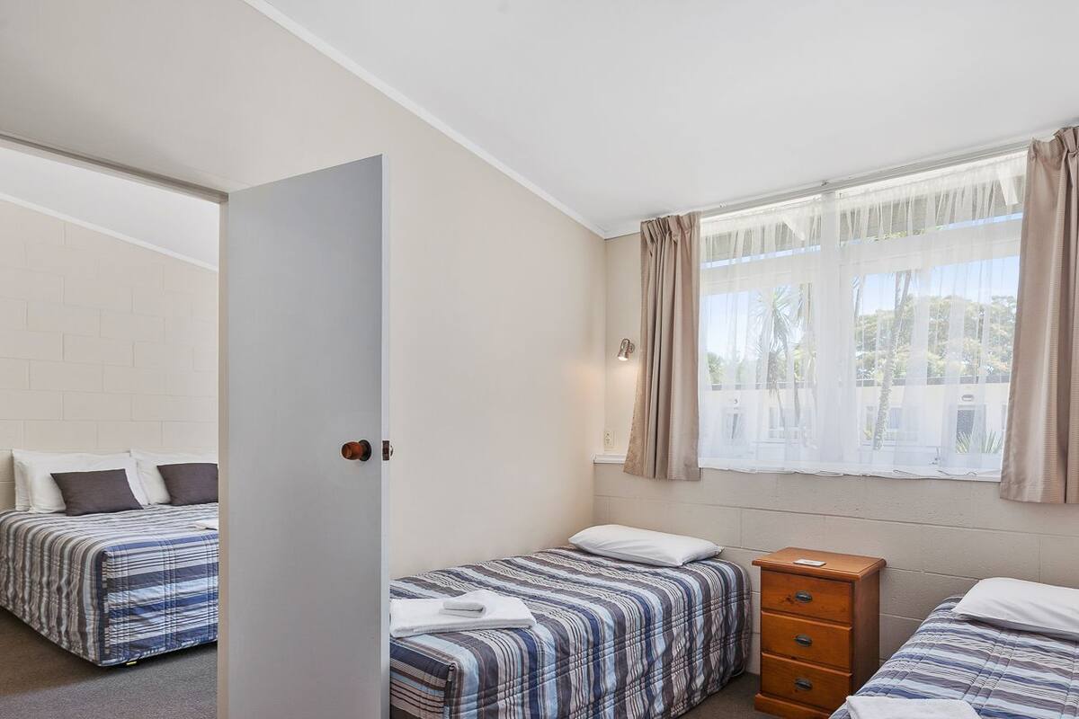 Een hotelkamer met twee bedden, een houten dressoir, een raam met dunne gordijnen, en een witte deur.