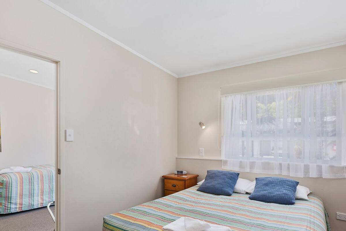 Een slaapkamer met een bed, nachtkastje, raam met dunne gordijnen en een wandlamp.