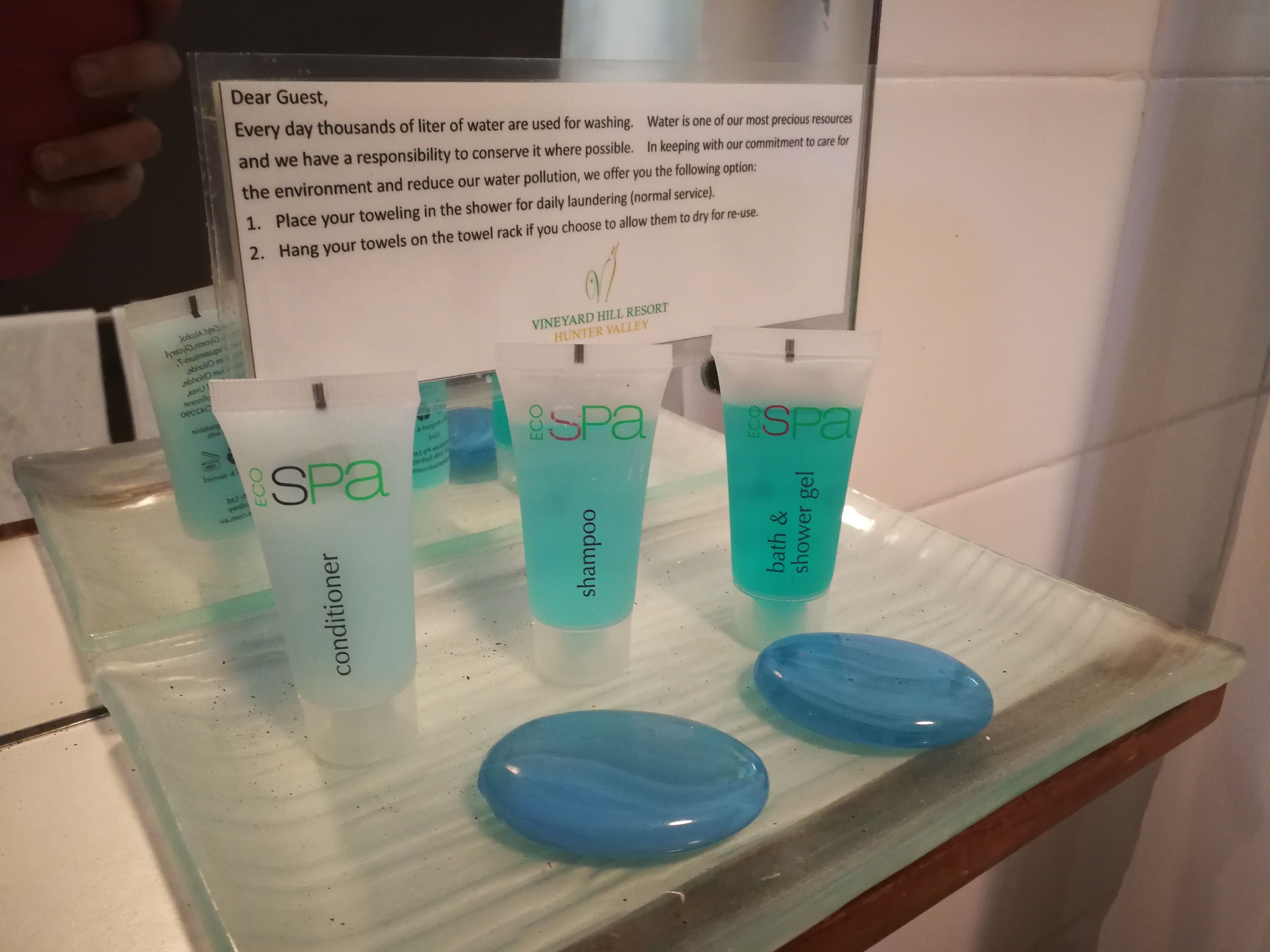 A glass display with four tubes of EcoSpa products: conditioner, shampoo, and bath & shower gel. A sign above explains water conservation efforts.