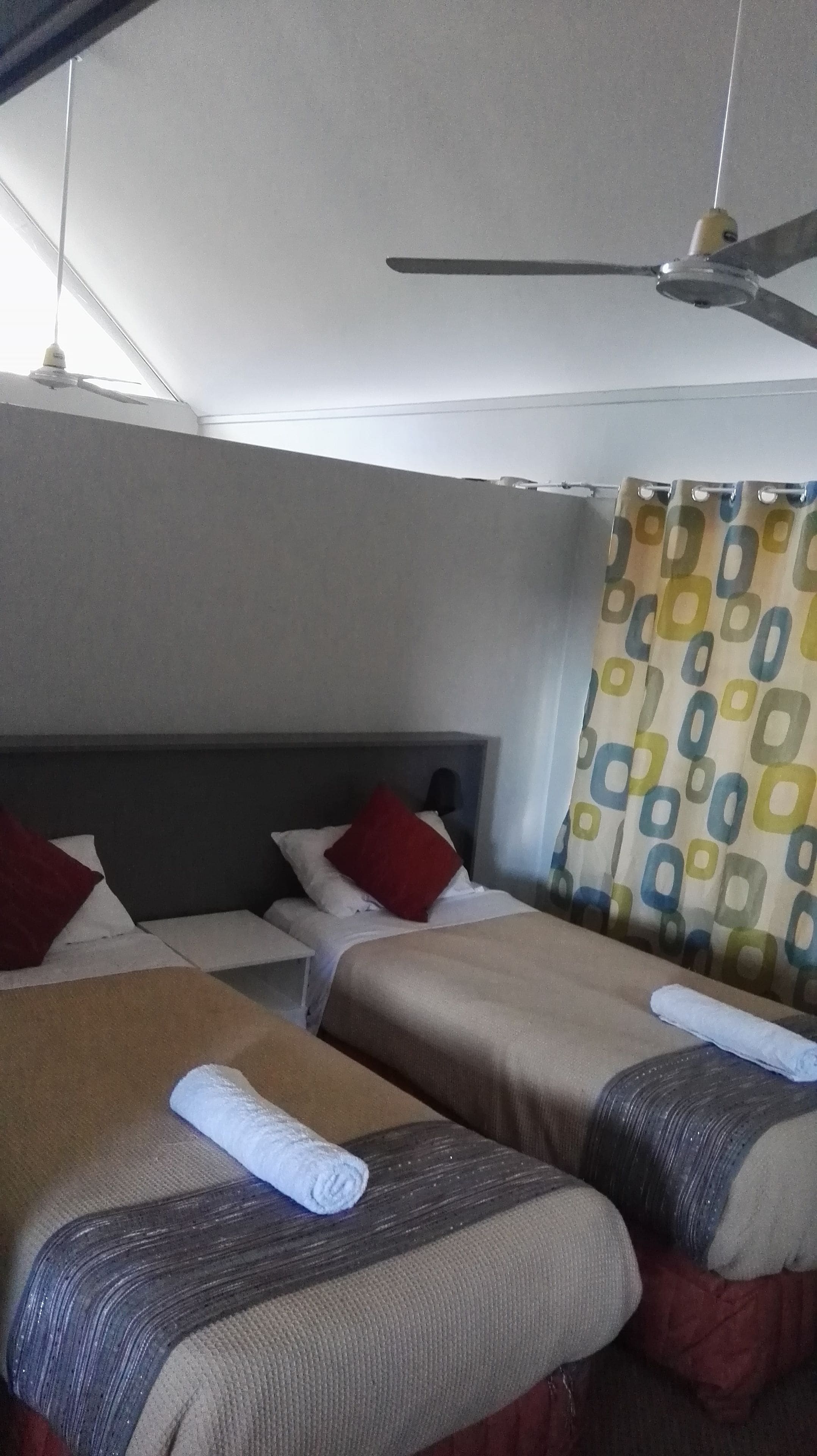 Hotelkamer met twee bedden, elk voorzien van een kussen en een opgerolde handdoek. Er is een plafondventilator en een gordijn met een patroon.