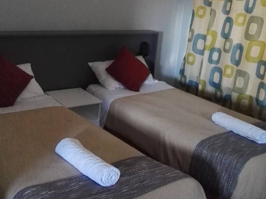 Habitación de hotel con dos camas, cada una con una almohada y una toalla enrollada. Hay un ventilador de techo y una cortina estampada.
