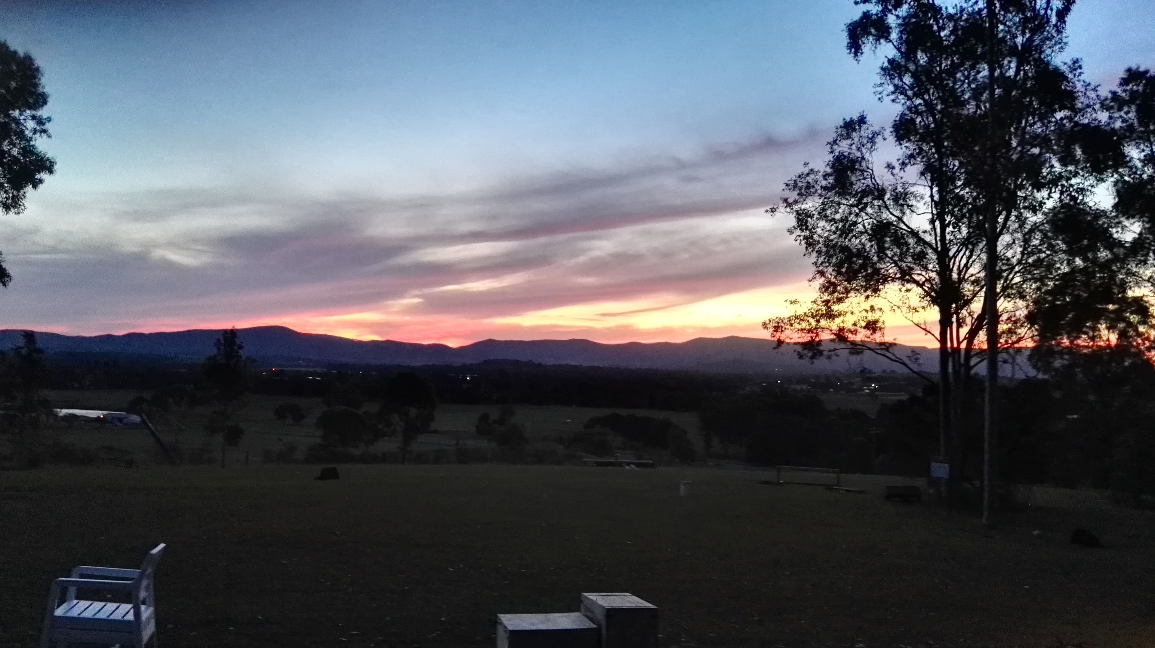 A sunset over a hilly landscape with a clear sky and a few trees in the foreground.
